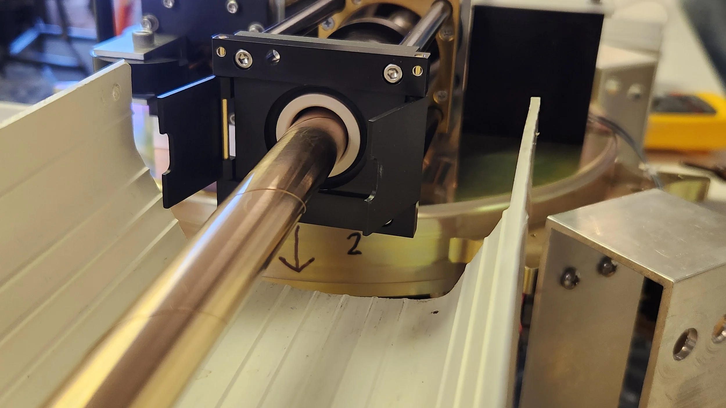 Close-up of a mechanical setup showing a metal rod passing through a black housing fixed on a gold-colored circular base. There are hand-drawn arrows and numbers on the base, indicating direction or measurements. The background includes other mechani
