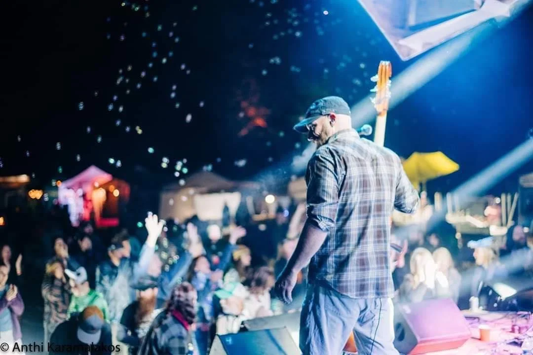 A man, Johnny Calamari,  on stage playing an upright bass at a festival, wearing a plaid shirt and a backward cap, facing an audience with raised hands under colorful stage lighting.