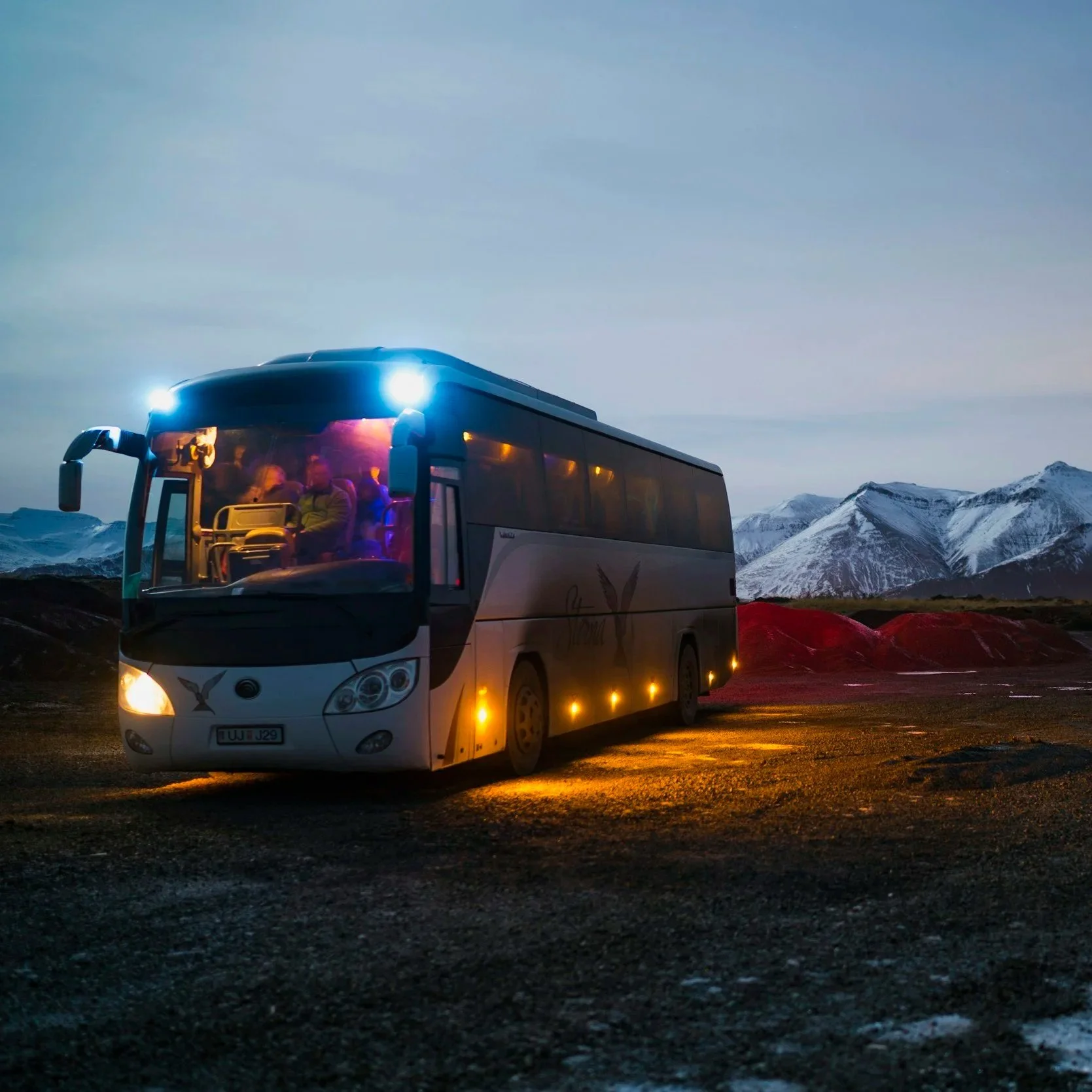 A tour bus parked outdoors during dusk with mountains in the background, some snow on the peaks, and landscape illuminated by the bus's lights.