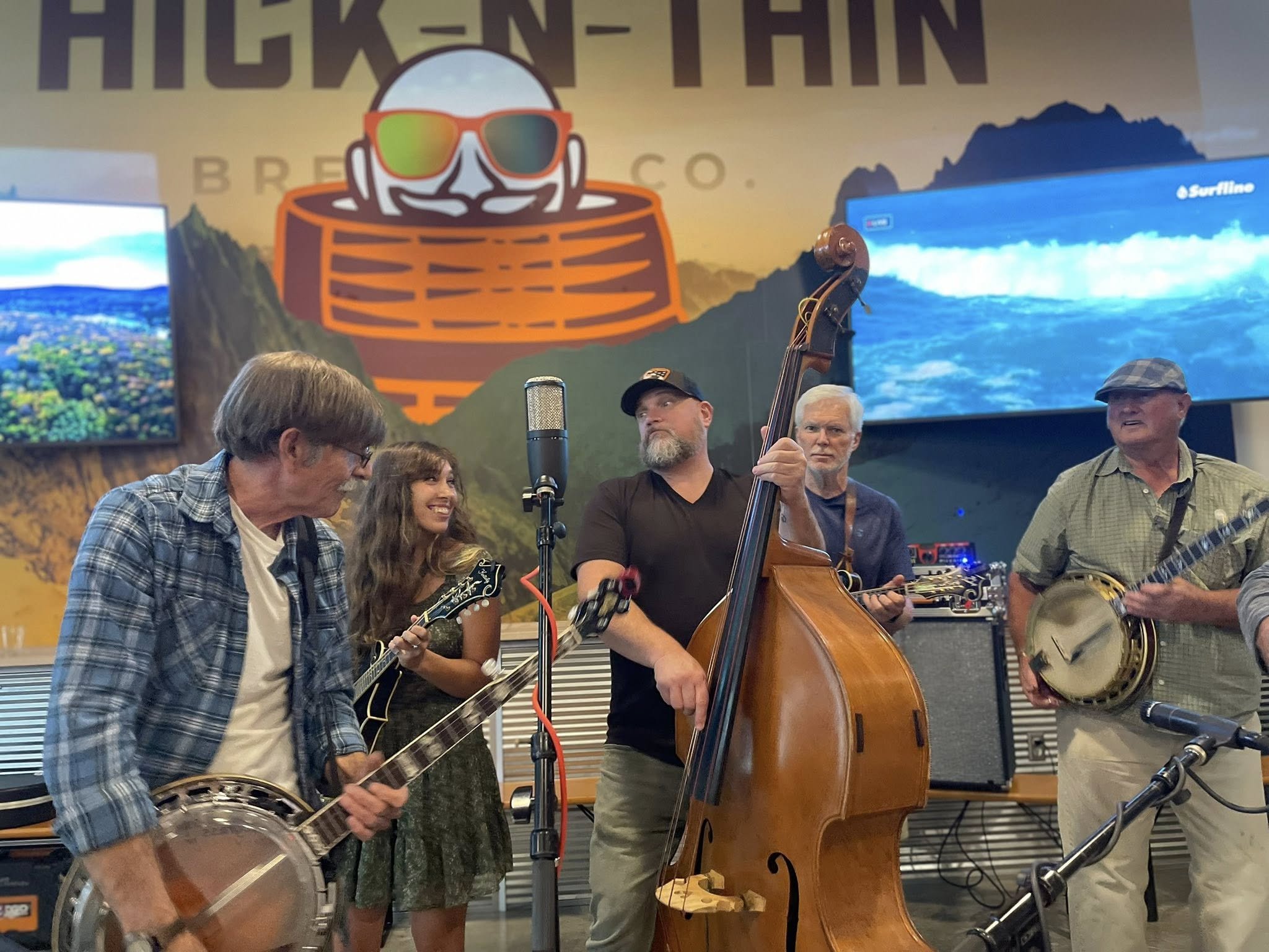 A group of five musicians, three men and one woman, playing instruments and performing together indoors. The background features a large colorful mural with the word 'HICKEN-THIN' and an illustration of a smiling face with sunglasses, mountains, and a television screen showing a wave.