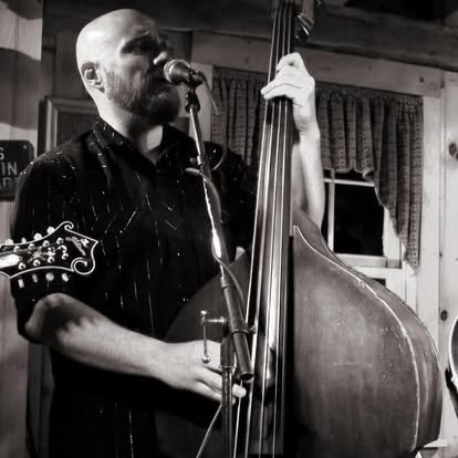 B man playing a double bass and singing into a microphone indoor with curtains in background.