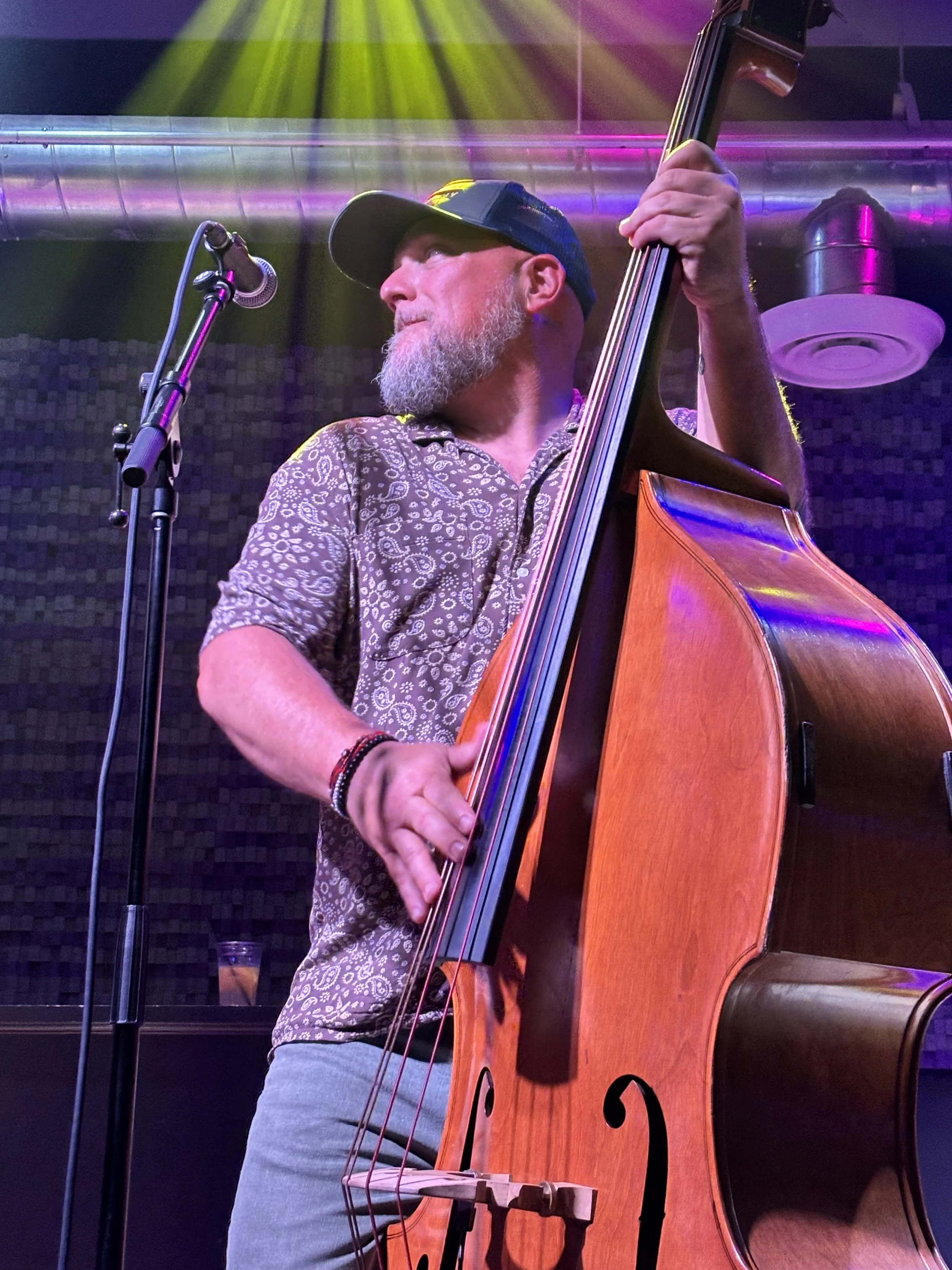 A man with a beard, wearing a baseball cap and a patterned shirt, playing an upright bass on stage with colorful stage lighting.