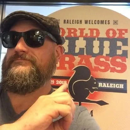 Man with sunglasses and a beard pointing at a poster that reads, "Raleigh welcomes World of Bluegrass" with a music note graphic.
