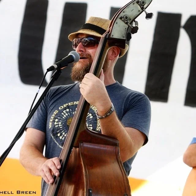 A man with a beard and sunglasses playing a double bass and singing into a microphone at an outdoor event.