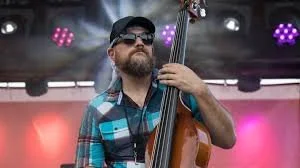 A man wearing sunglasses and a baseball cap playing a double bass on stage with colorful lights.