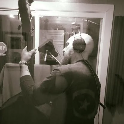 A man with headphones recording vocals in a studio, holding a microphone close to his mouth.