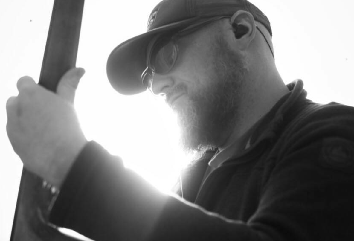 A man wearing a hat and sunglasses looking down at a tablet or similar device.