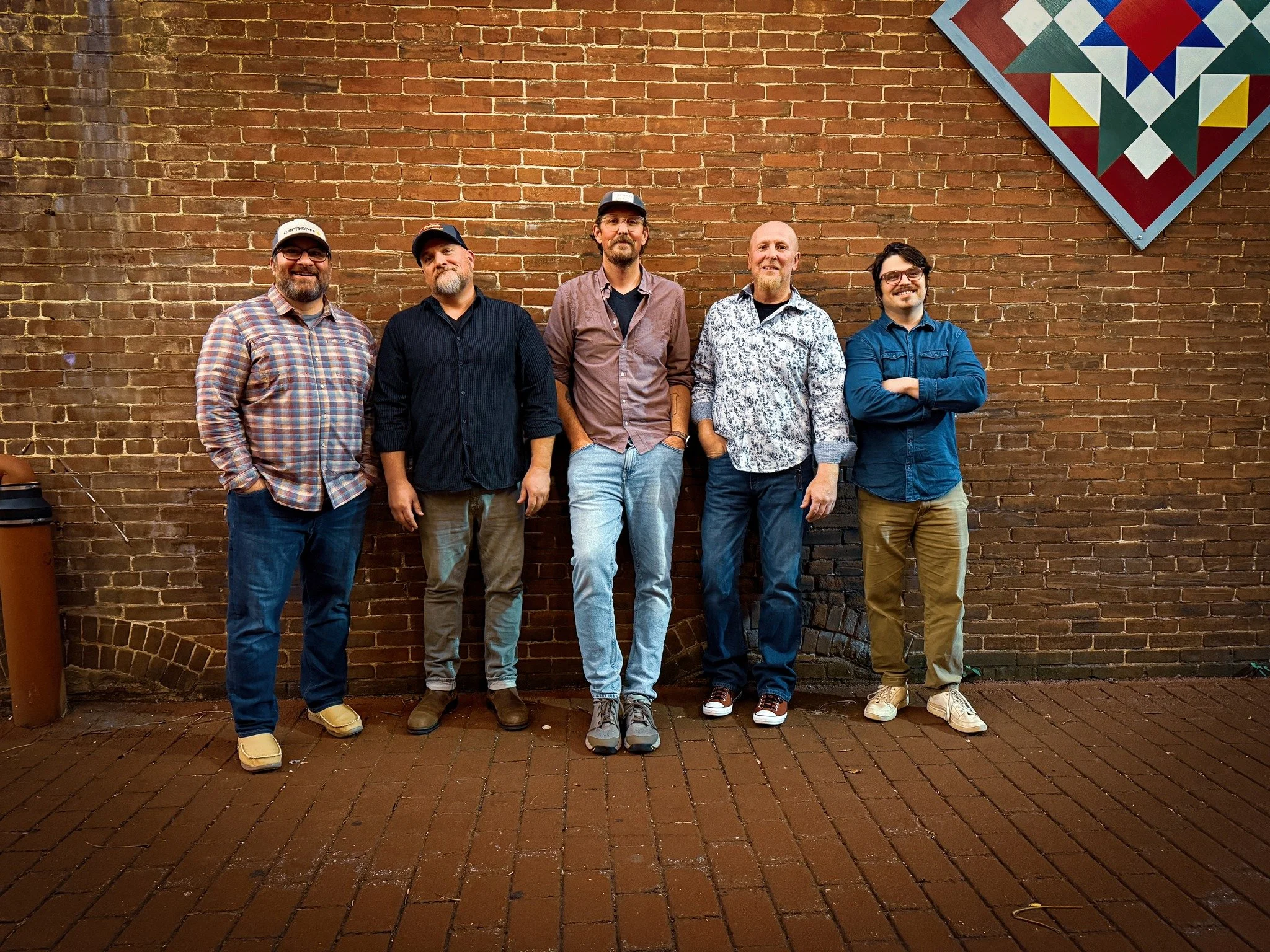 Group of five men standing against a brick wall, some with arms crossed or hands in pockets, smiling, with a quilt hanging on the wall in the top right corner.
