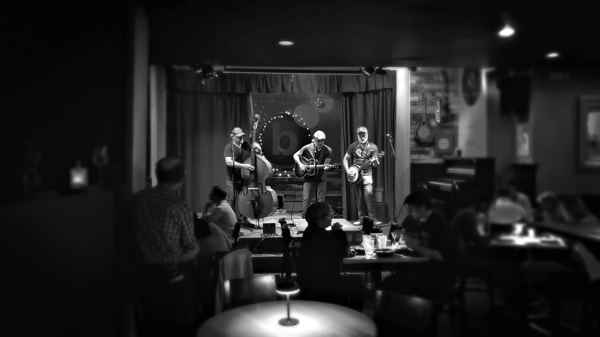 A live music performance shows three musicians playing guitars on a small stage with a curtain backdrop. The audience sits at tables in a dimly lit venue, with some patrons watching the performance.