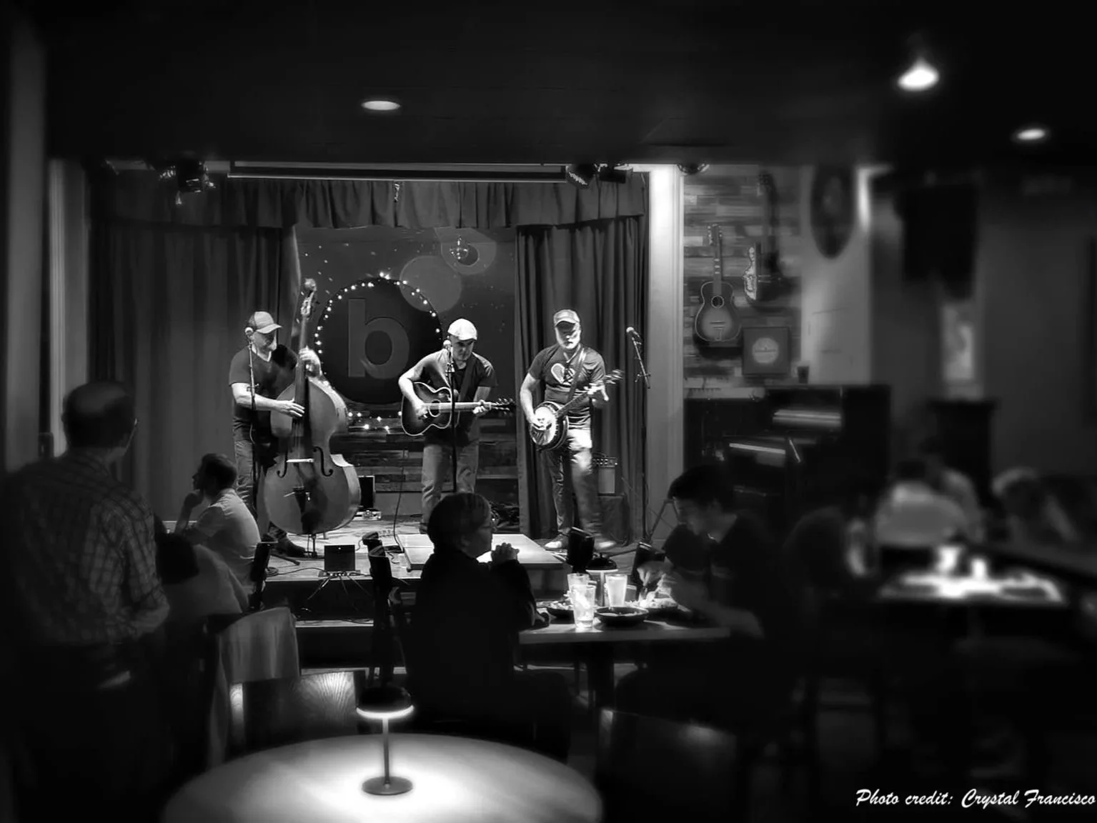 A black and white photo of a live music performance at a small venue, with three musicians playing acoustic instruments on stage and an audience seated at tables in the foreground.