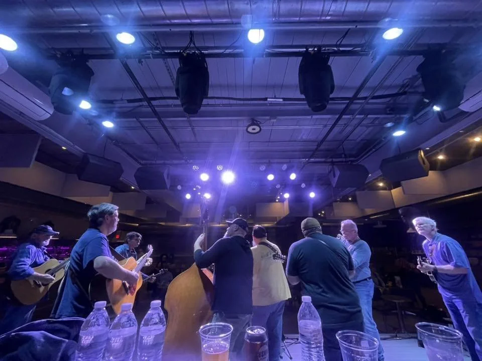 A group of musicians playing guitars on stage under blue stage lights in a concert hall, with water bottles and cups placed on a table in the foreground.