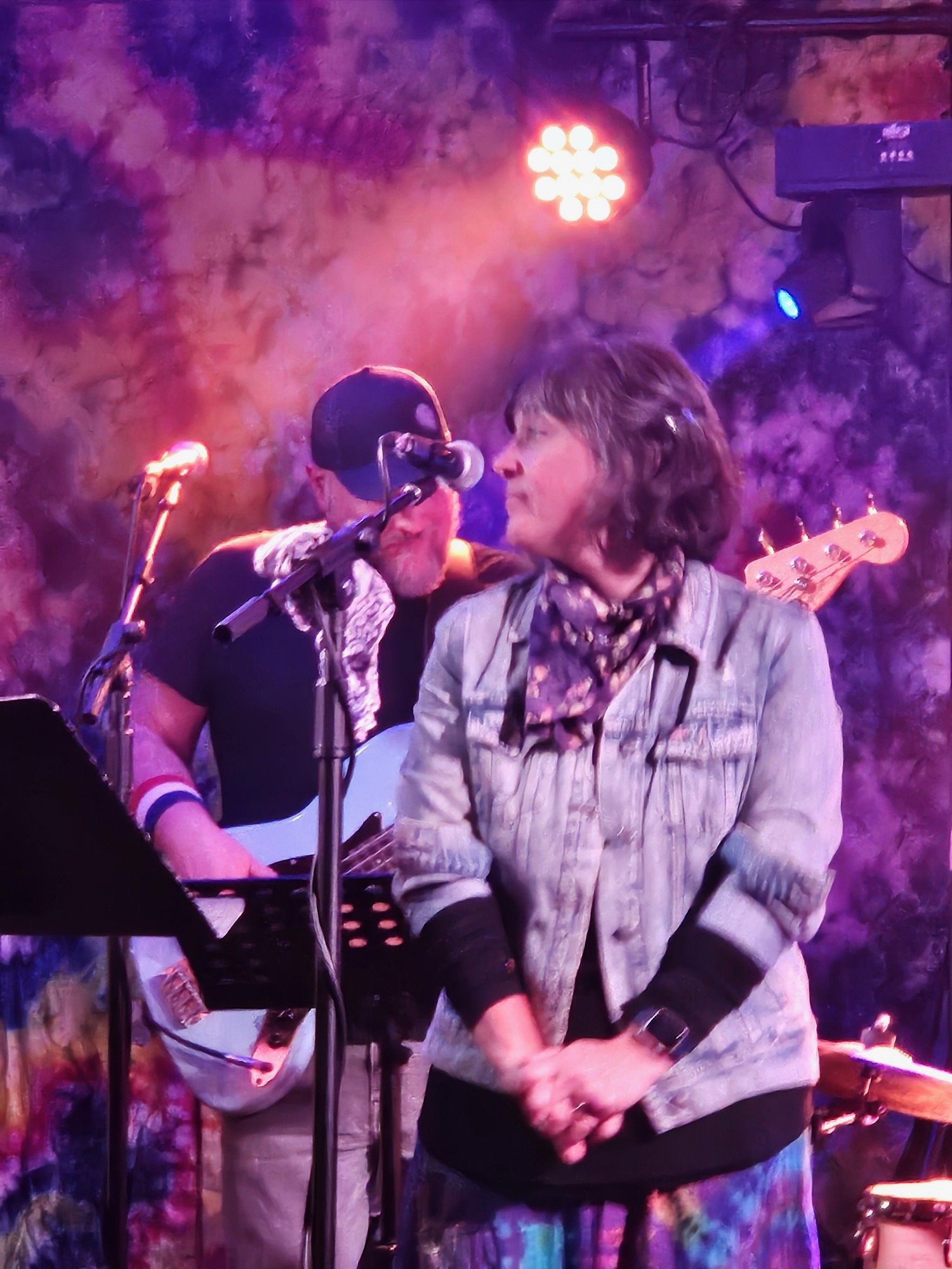 A woman with short hair in a light jacket and scarf stands on stage with her hands clasped in front of her, as a man with a beard, glasses, and a cap behind her sings into a microphone. The background is colorful with purple and pink hues, with stage