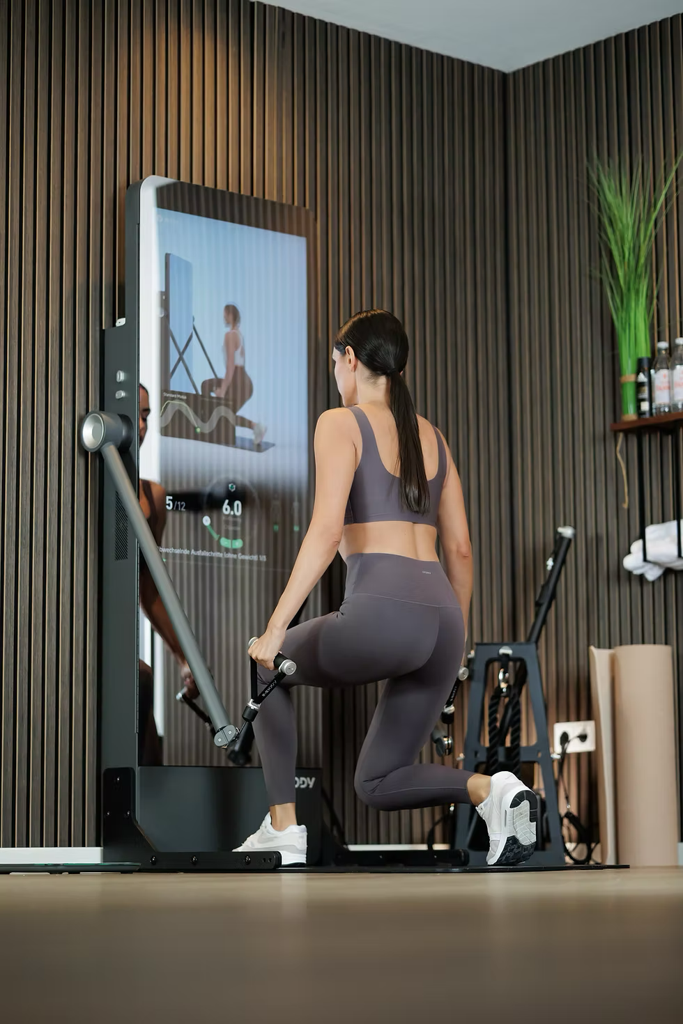 A woman is exercising on a rowing machine in a gym, facing a mirror with a digital display showing her workout progress.