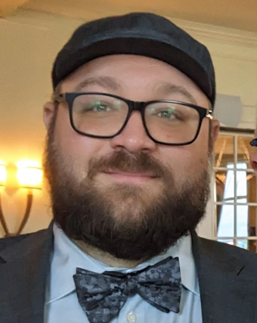 A man wearing glasses, a black hat, a light blue dress shirt, and a patterned black bow tie. Background includes warm lighting and part of a window with curtains.