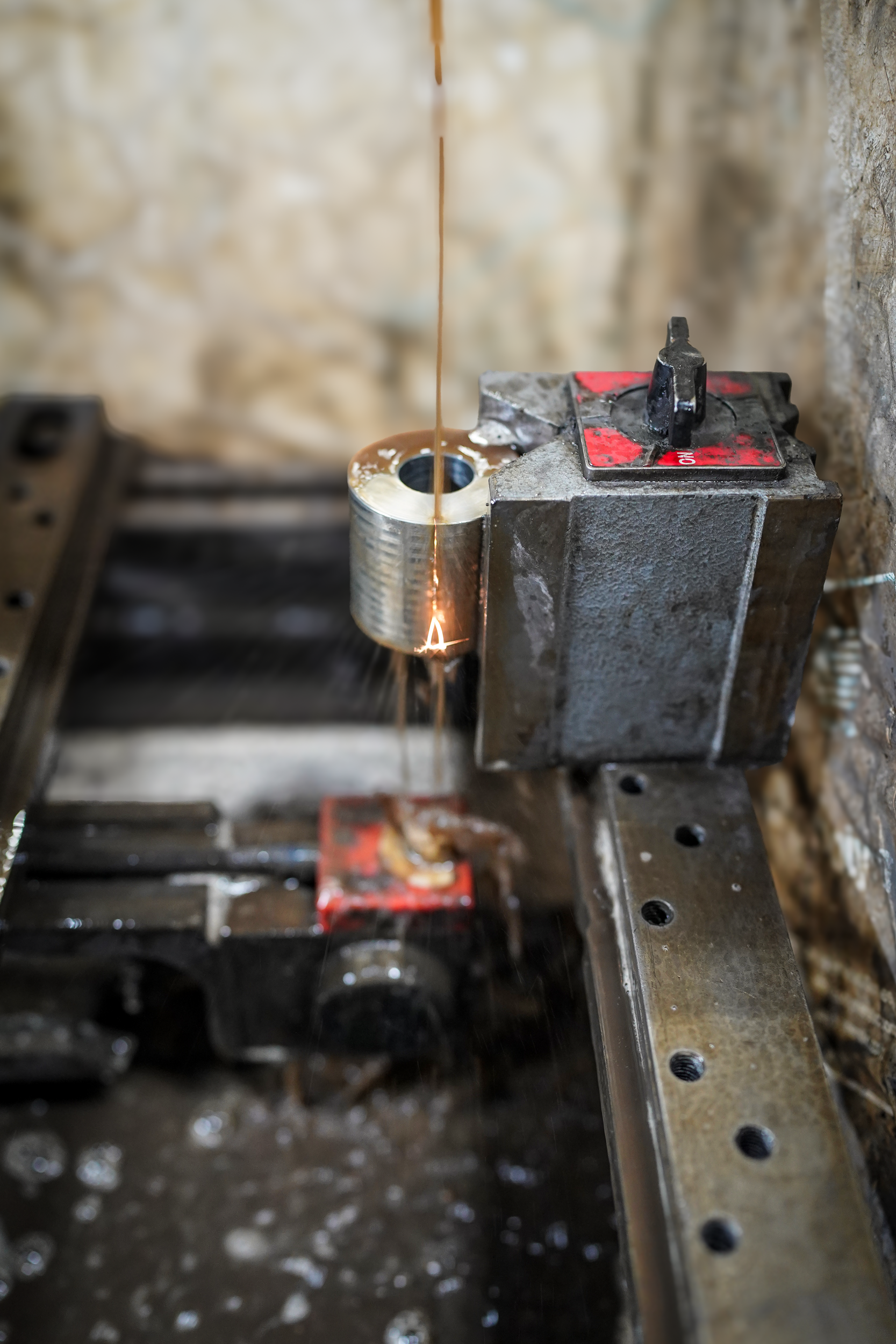 A metalworking or welding process with sparks and molten metal, involving a machine with a lever and a circular metal piece, set in a workshop with stone walls.