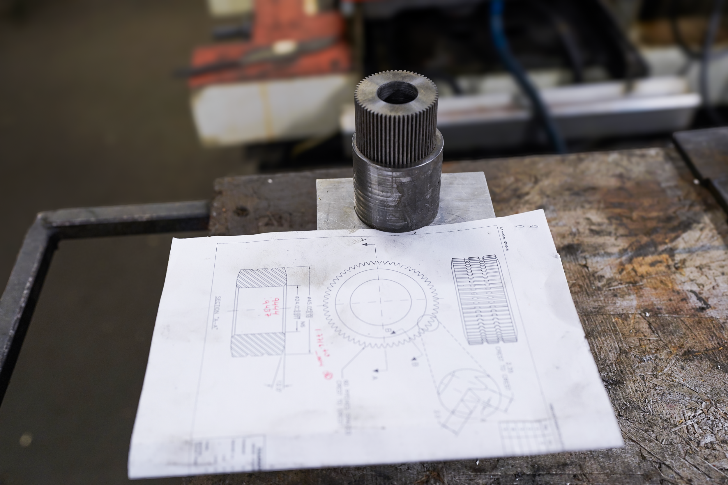 Metal machine gear shaft placed on a technical drawing sheet on a wooden workbench in a workshop.