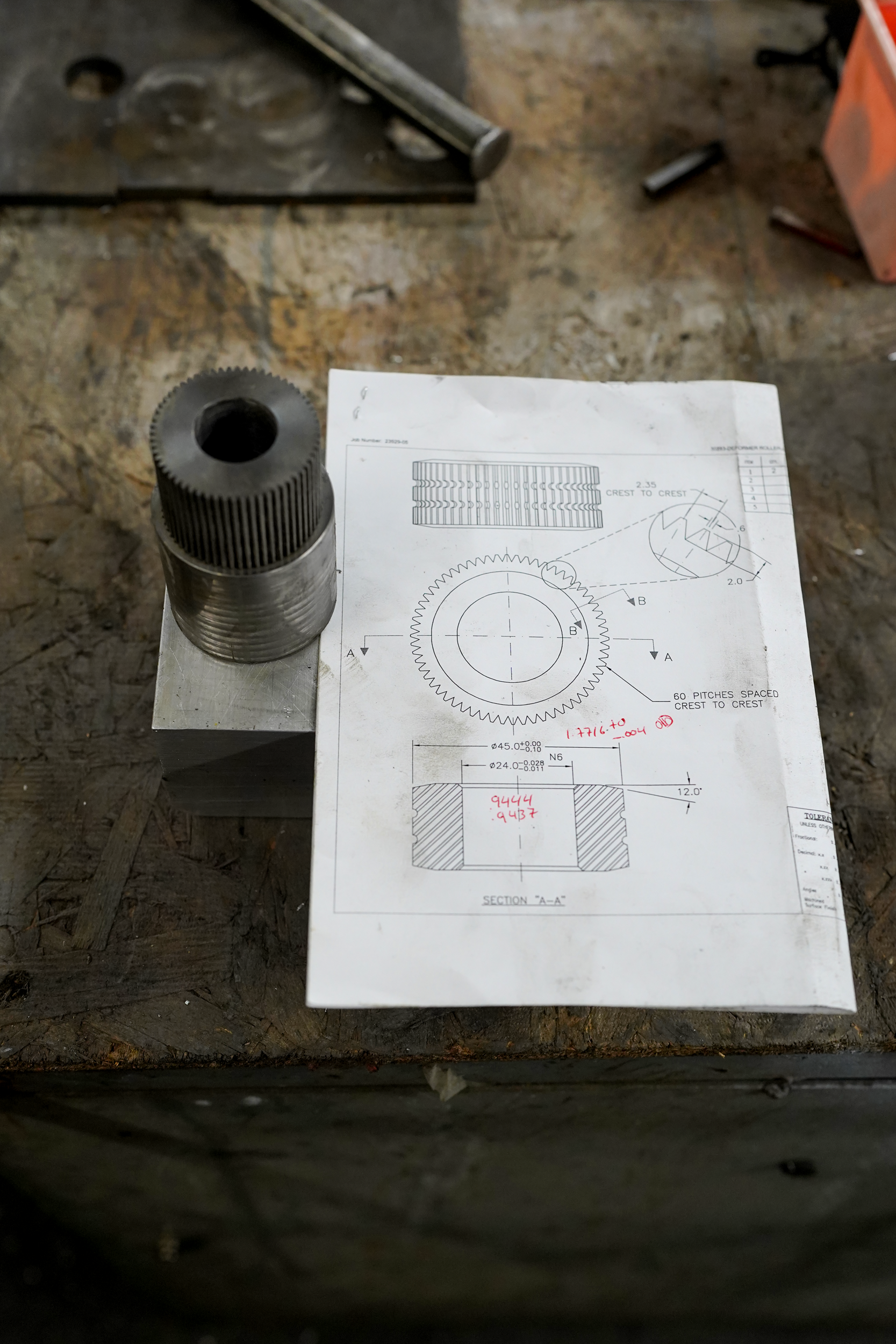 A metal gear part sitting on top of a technical drawing with detailed measurements and specifications, placed on a workbench in a workshop setting.