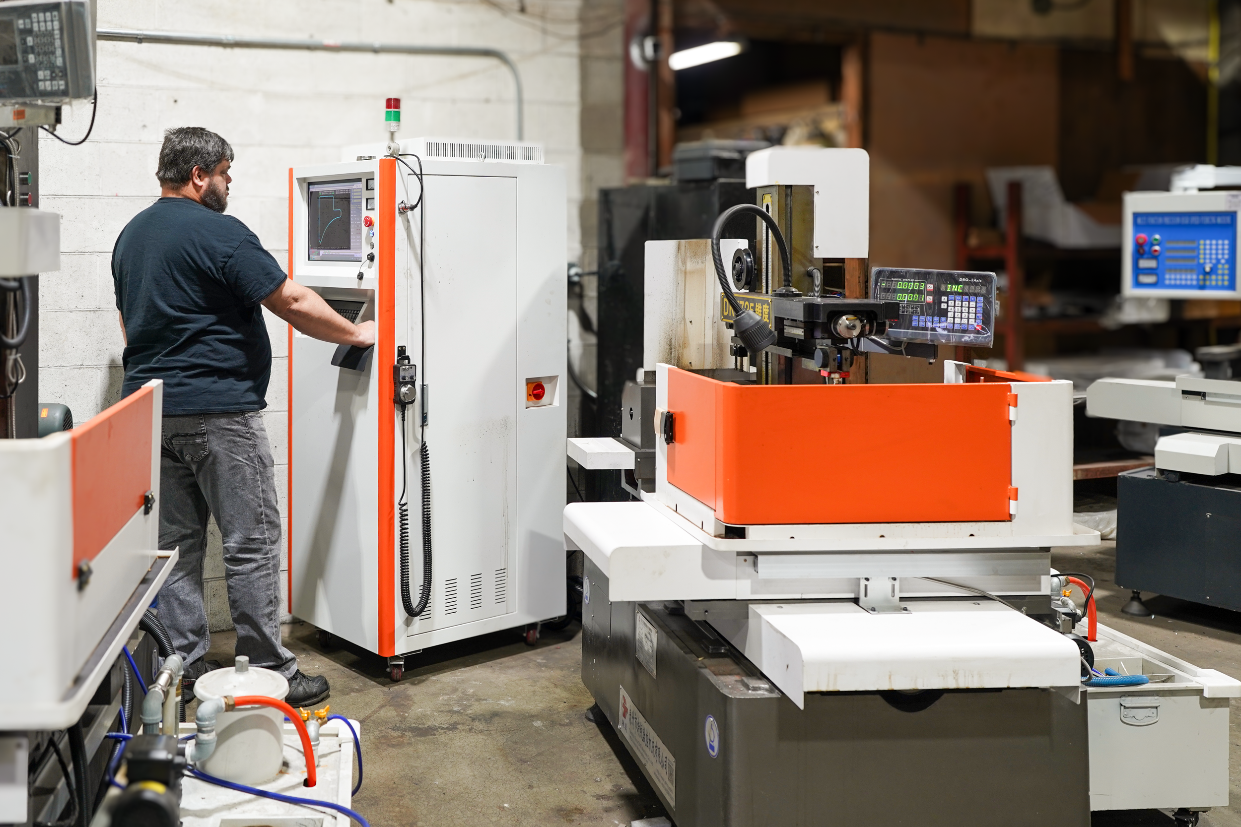 A man operating a CNC machine in an industrial workshop with shelves and equipment in the background.
