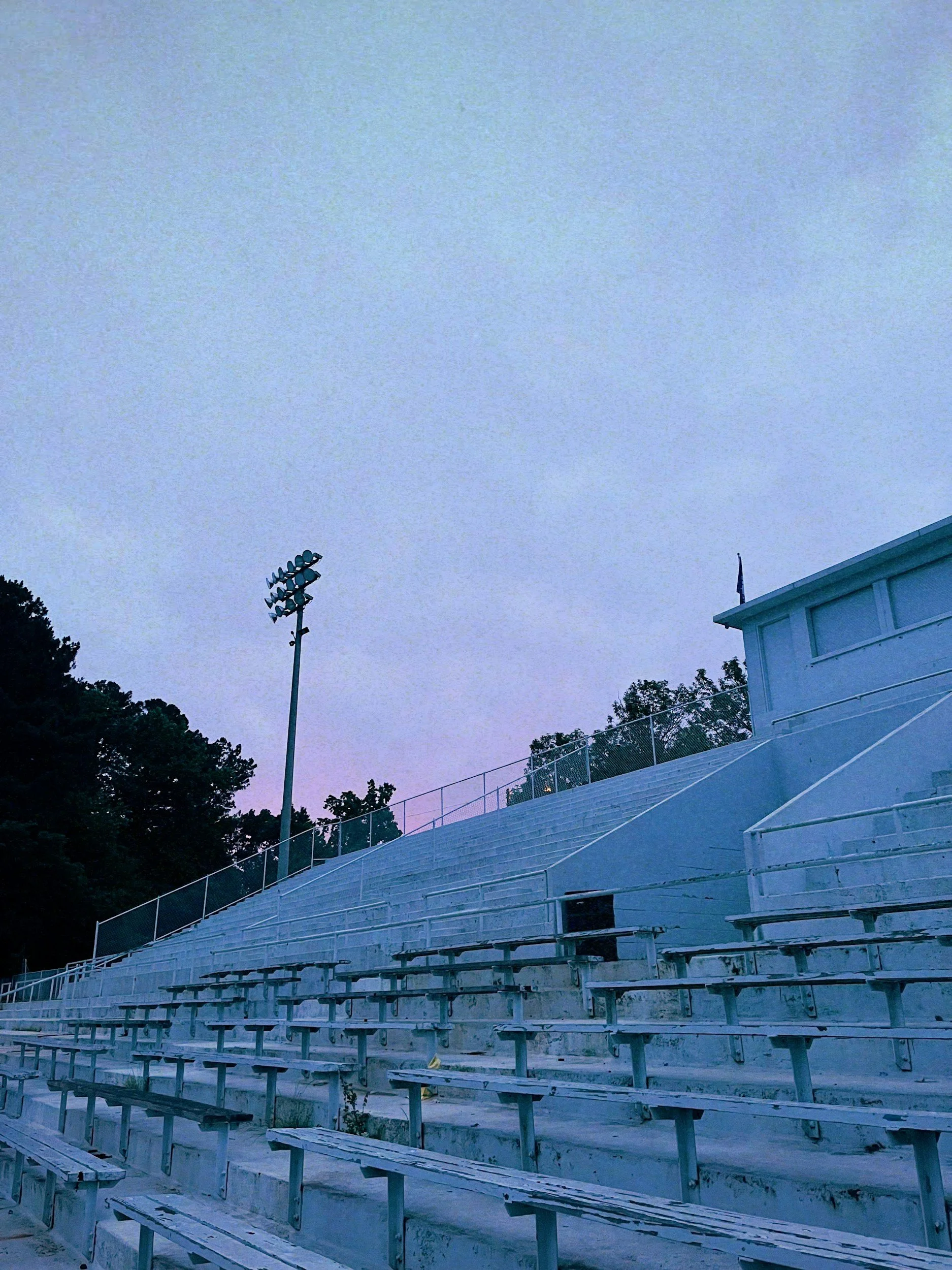 Empty outdoor football stadium bleachers with a light pole and trees in the background under a cloudy sky.