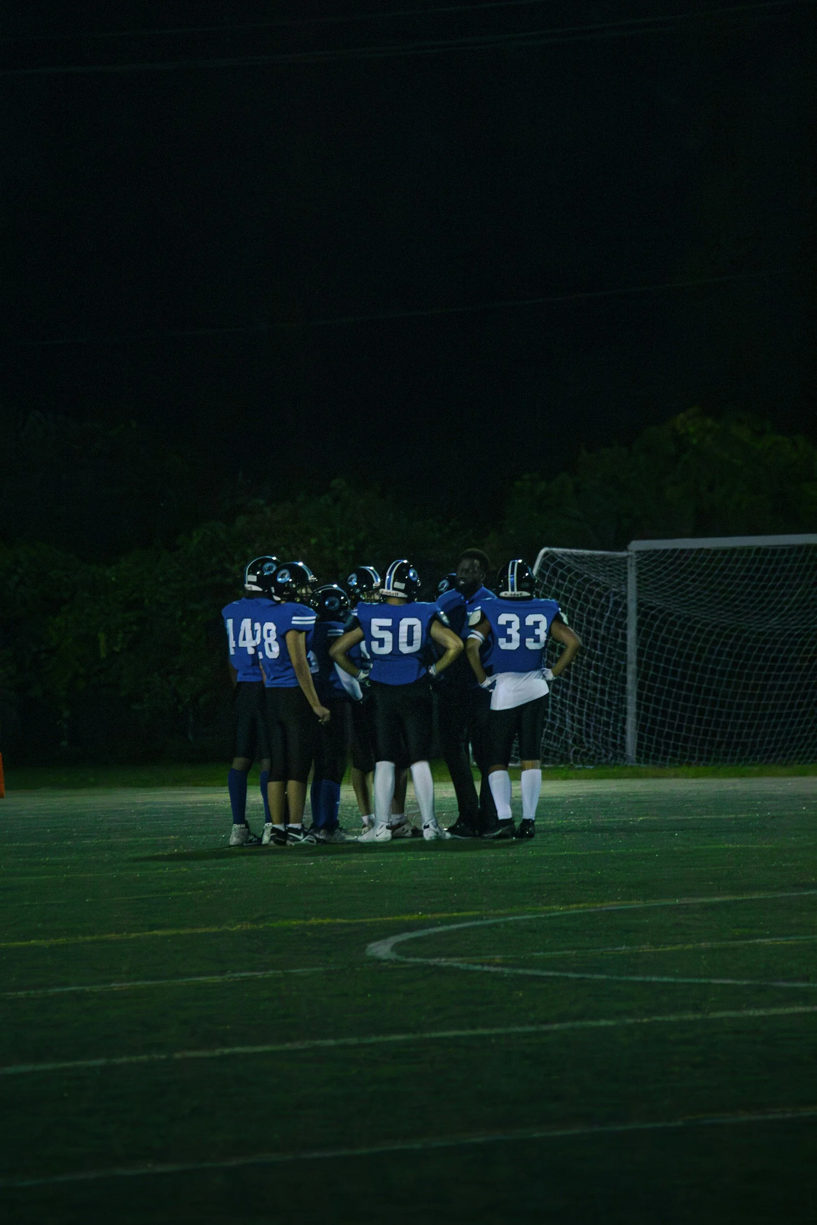 A group of football players in blue jerseys huddle together on a football field at night.