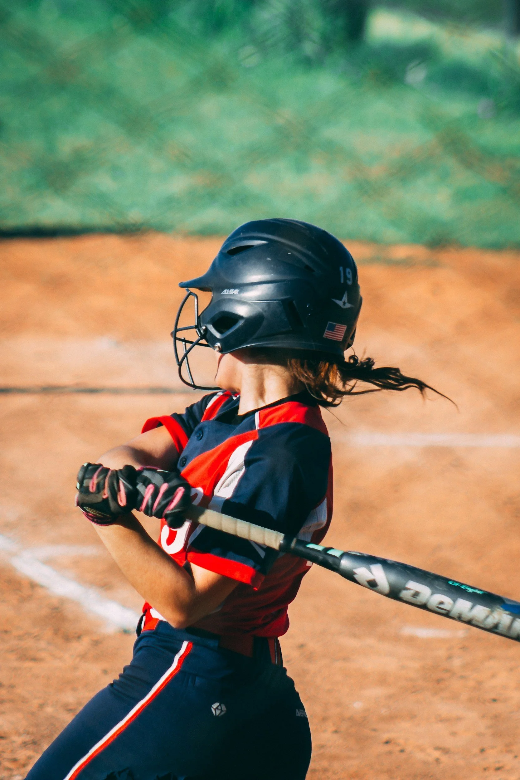 A female softball player wearing a black helmet, red and navy blue uniform, and gloves, swinging a bat on the field.