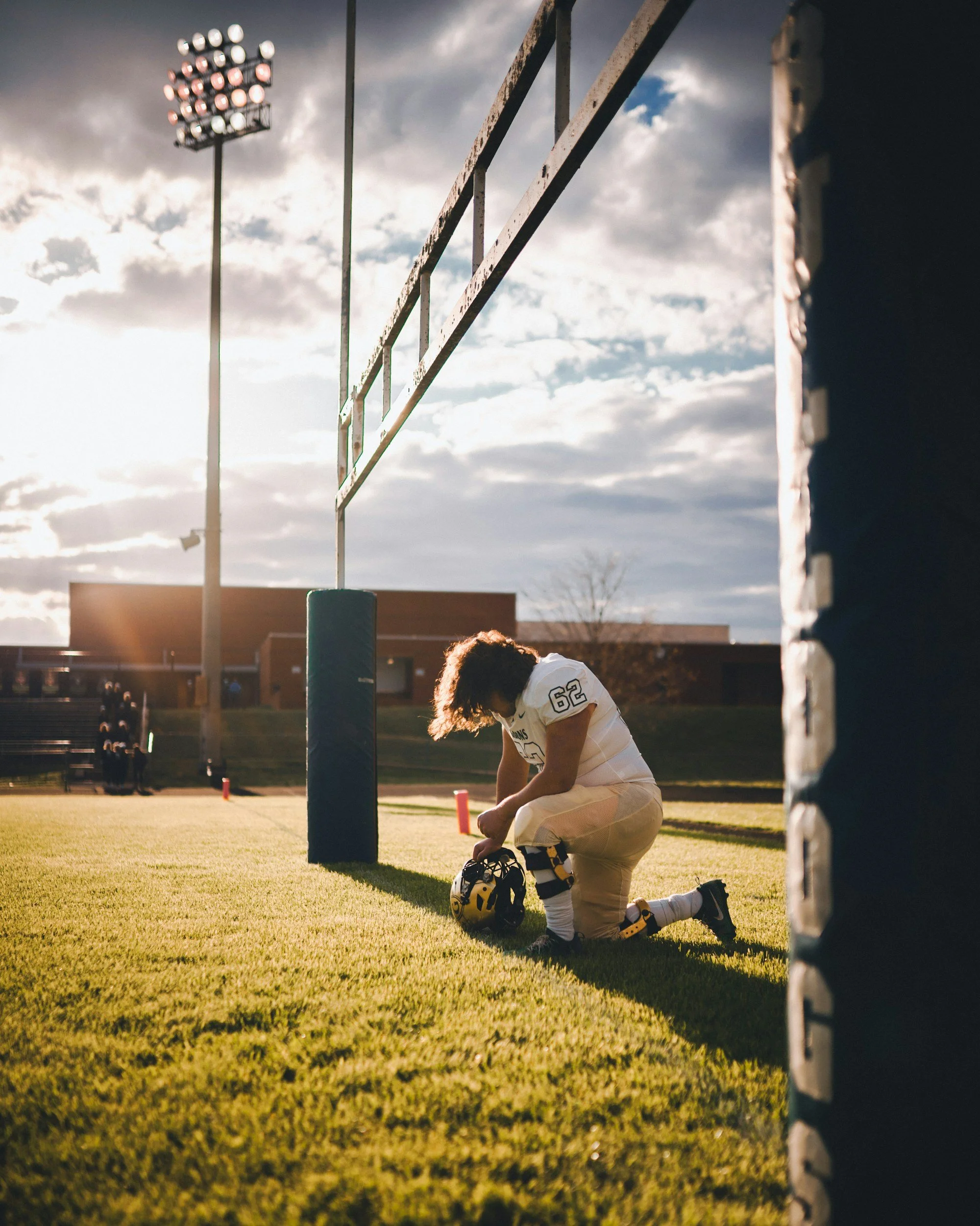 A football player kneeling and praying on the field during sunset, adjusting his helmet near the goalpost on a well-lit football field.
