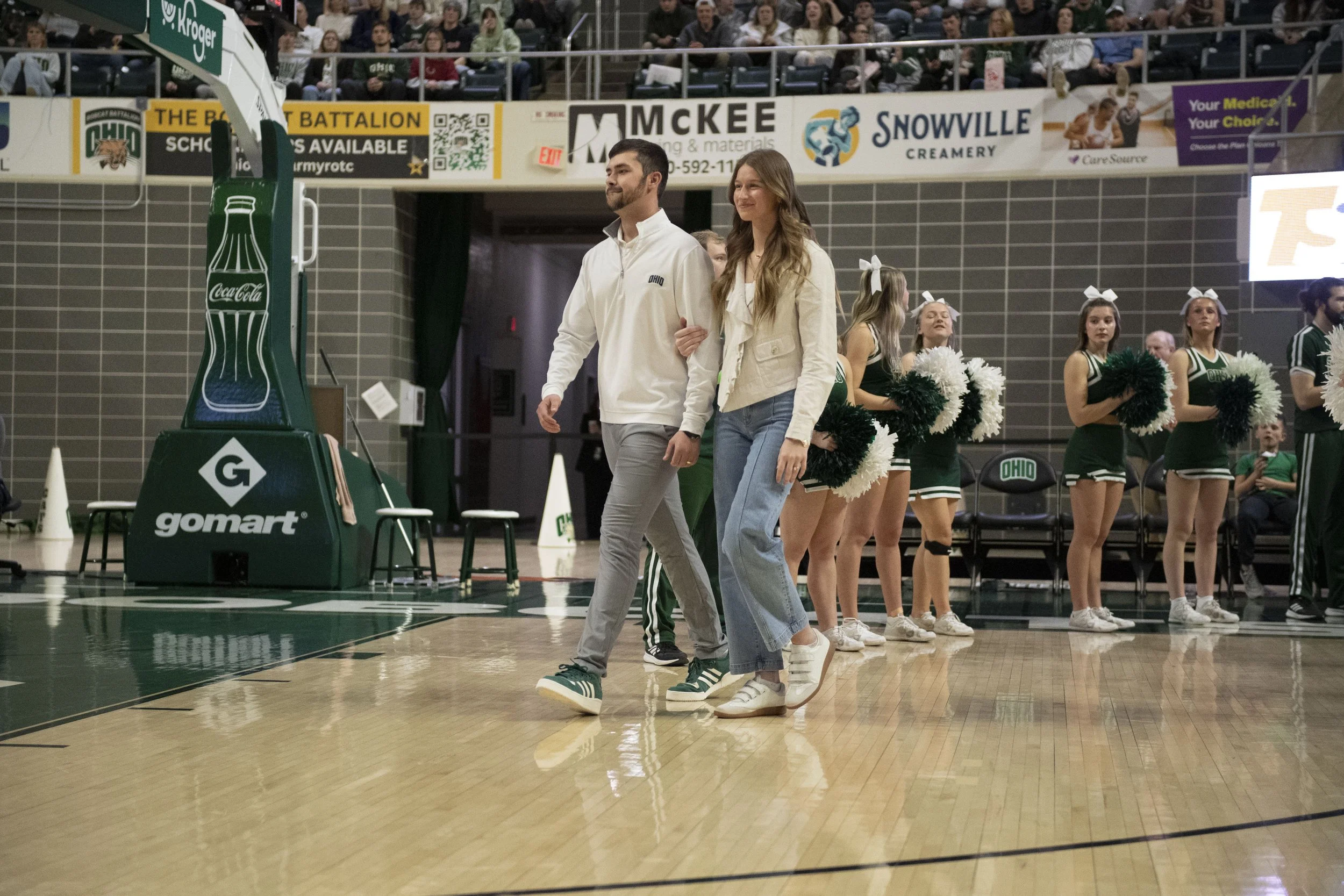 Brooks and Chariti McKeller walking onto a basketball court, surrounded by cheerleaders holding pom-poms, with spectators watching from the stands.