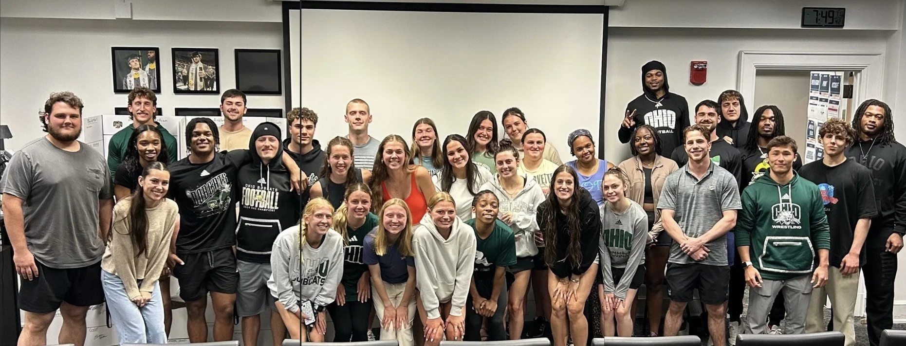 A large group of young adults posing together in a classroom or meeting room, smiling at the camera with some making peace signs.