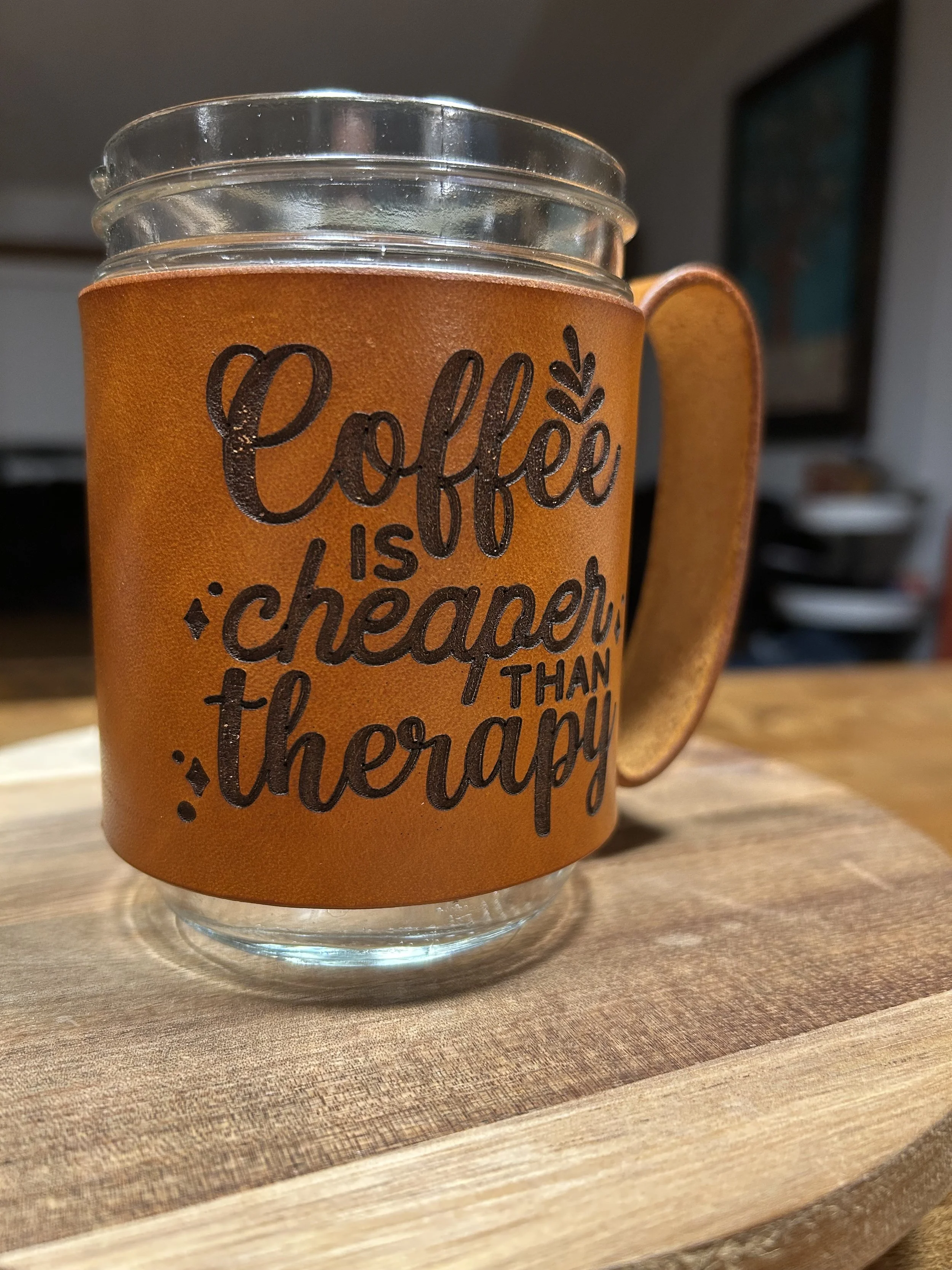 Close-up of a mason jar mug with a leather sleeve that has black text reading "Coffee is better than therapy" resting on a wooden surface in a room with a blurred background.