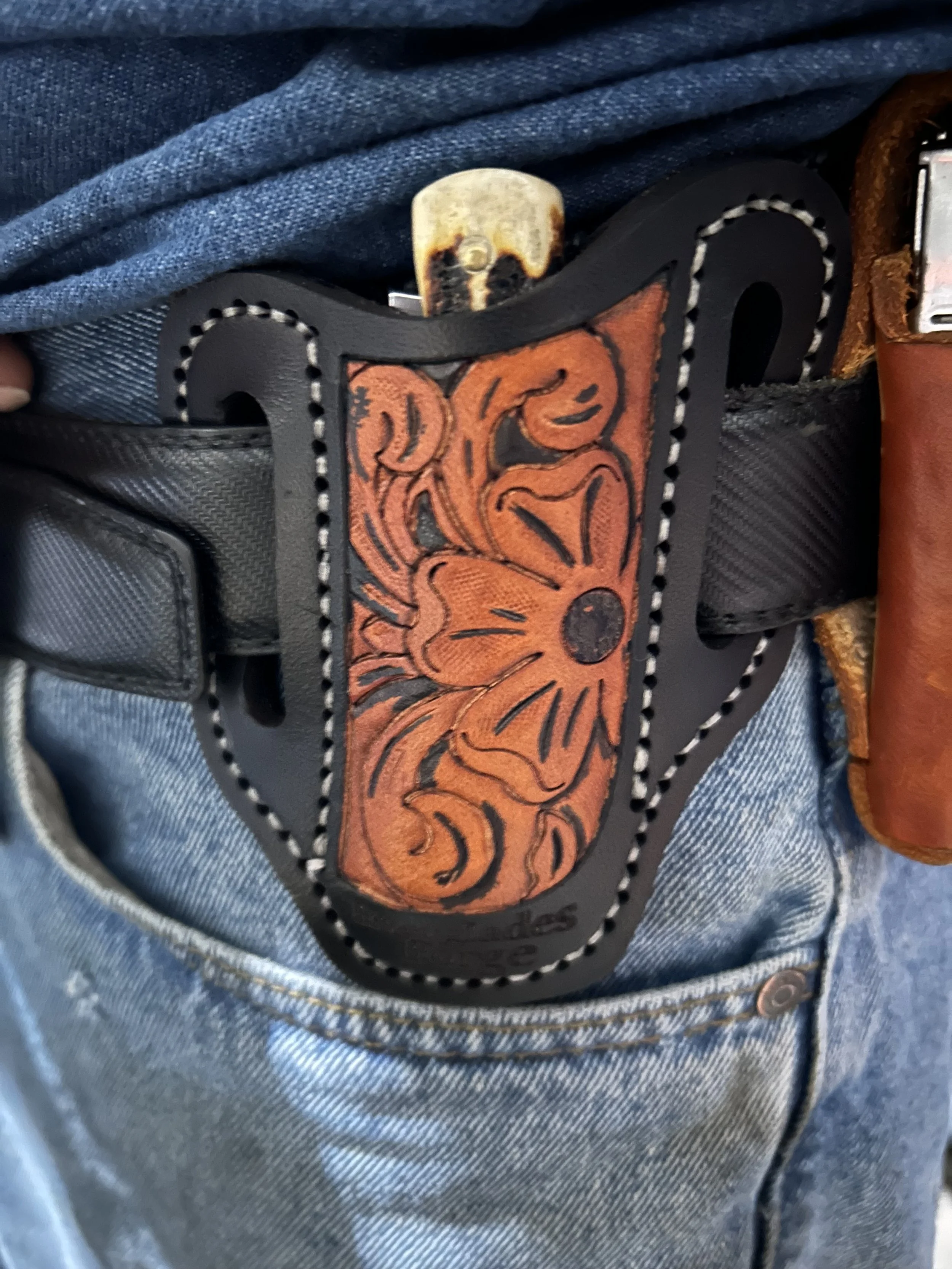 Close-up of a leather pocket knife holster/sheath with orange floral design, attached to blue denim jeans with a leather loop and buckle.