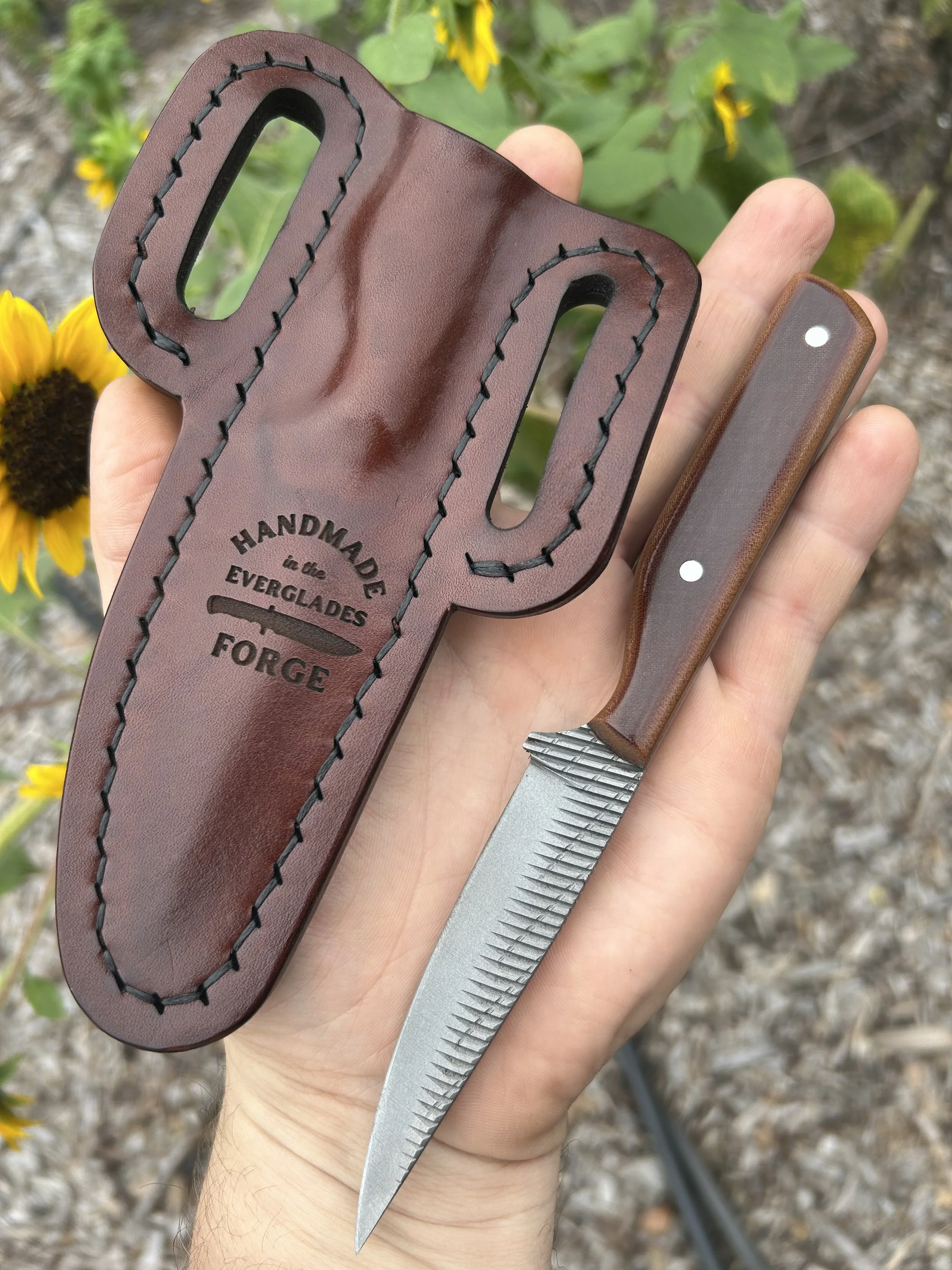 A hand holding a leather knife sheath with stitching, a fixed-blade knife with a brown micarta handle, and a small section of yellow flowers in the background.