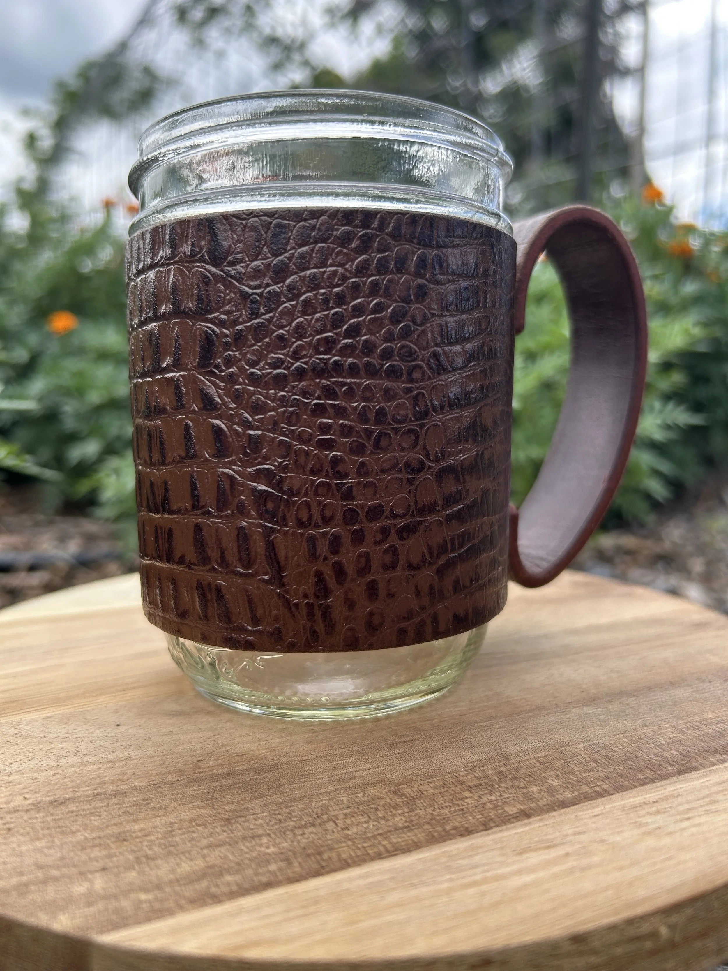 Close-up of a glass mug with a faux alligator leather sleeve, placed on a wooden surface outdoors with greenery and cloudy sky in the background.