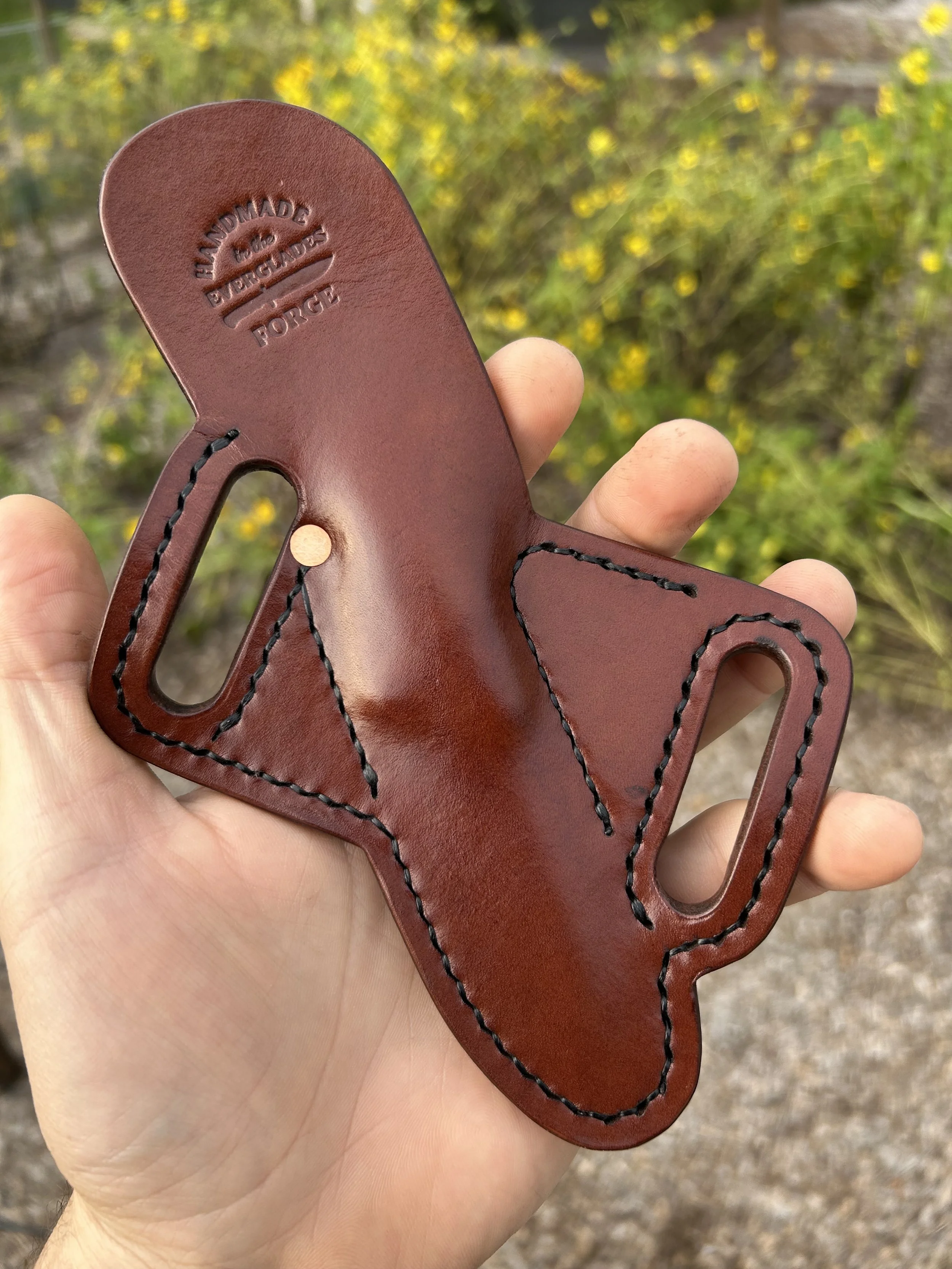 Close-up of a leather knife sheath with black stitching, held in a person's hand, with a background of yellow flowers.