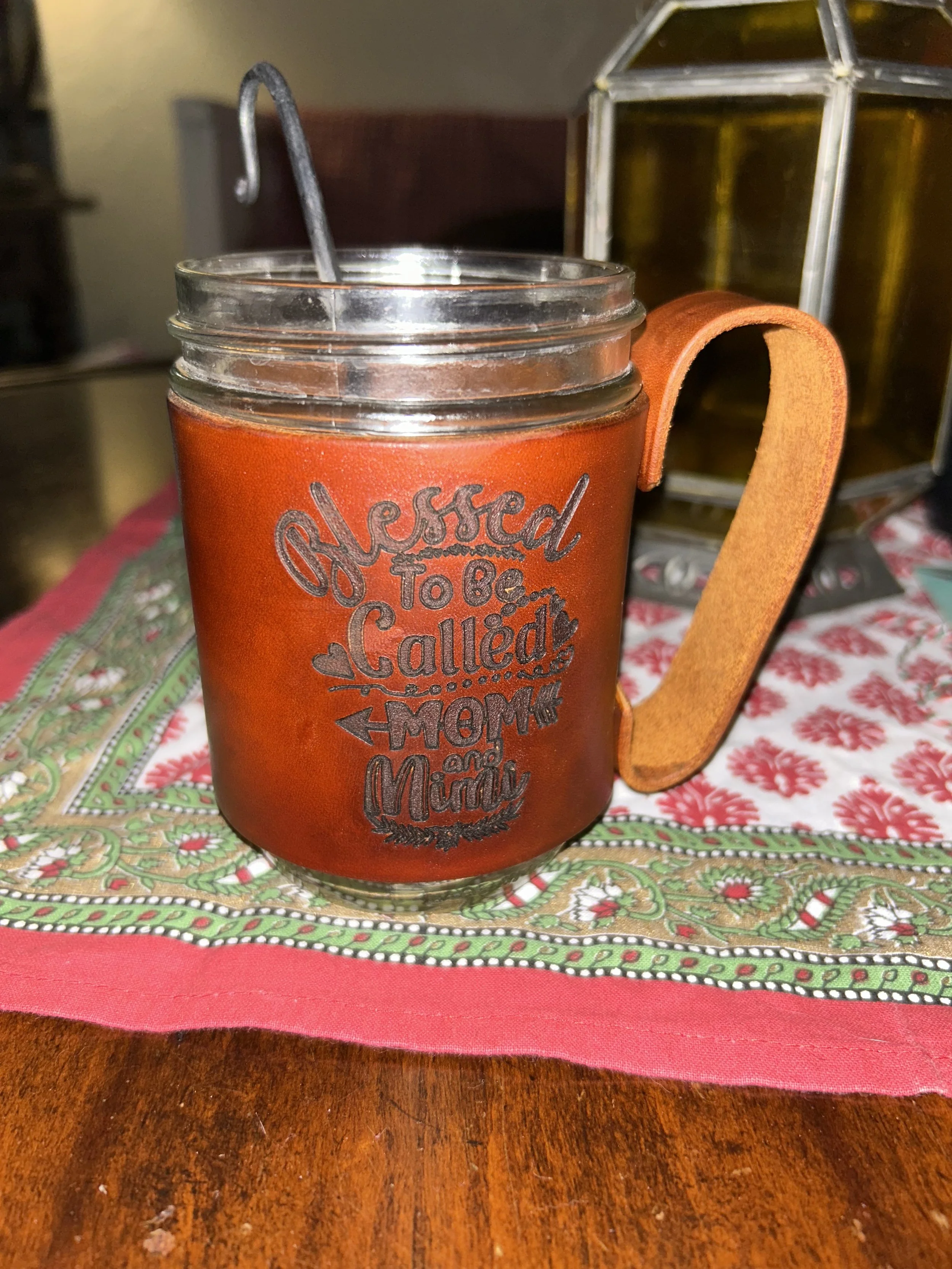 A glass mason jar with a leather sleeve that has the phrase 'Blessed to be called Mom and Mummy' written on it, placed on a decorative tablecloth on a wooden table. The jar has a metal straw inside.