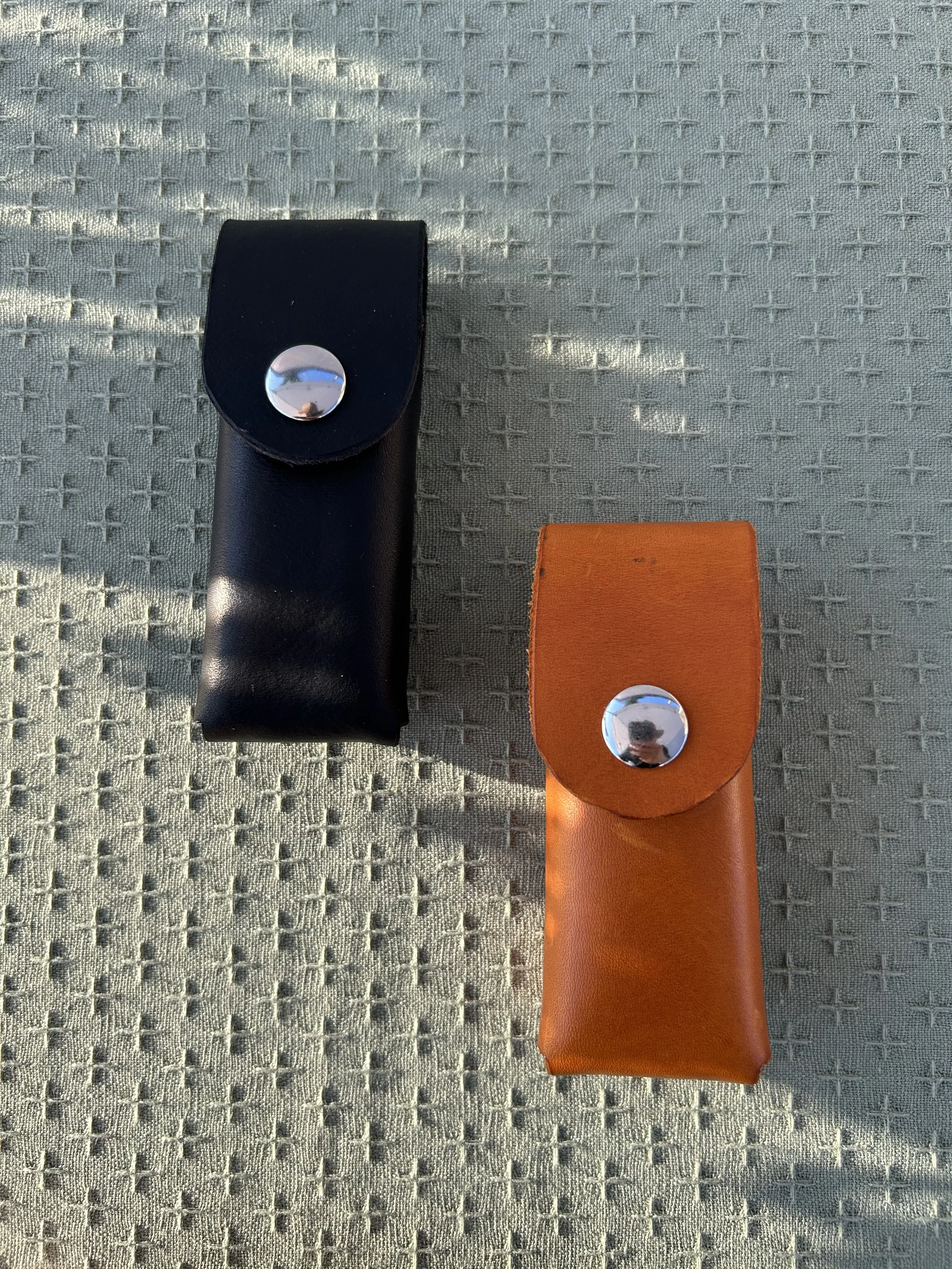 Close-up of two leather multitool knife sheaths, one black and one orange, each with a shiny metallic snap button, placed on a textured fabric surface.