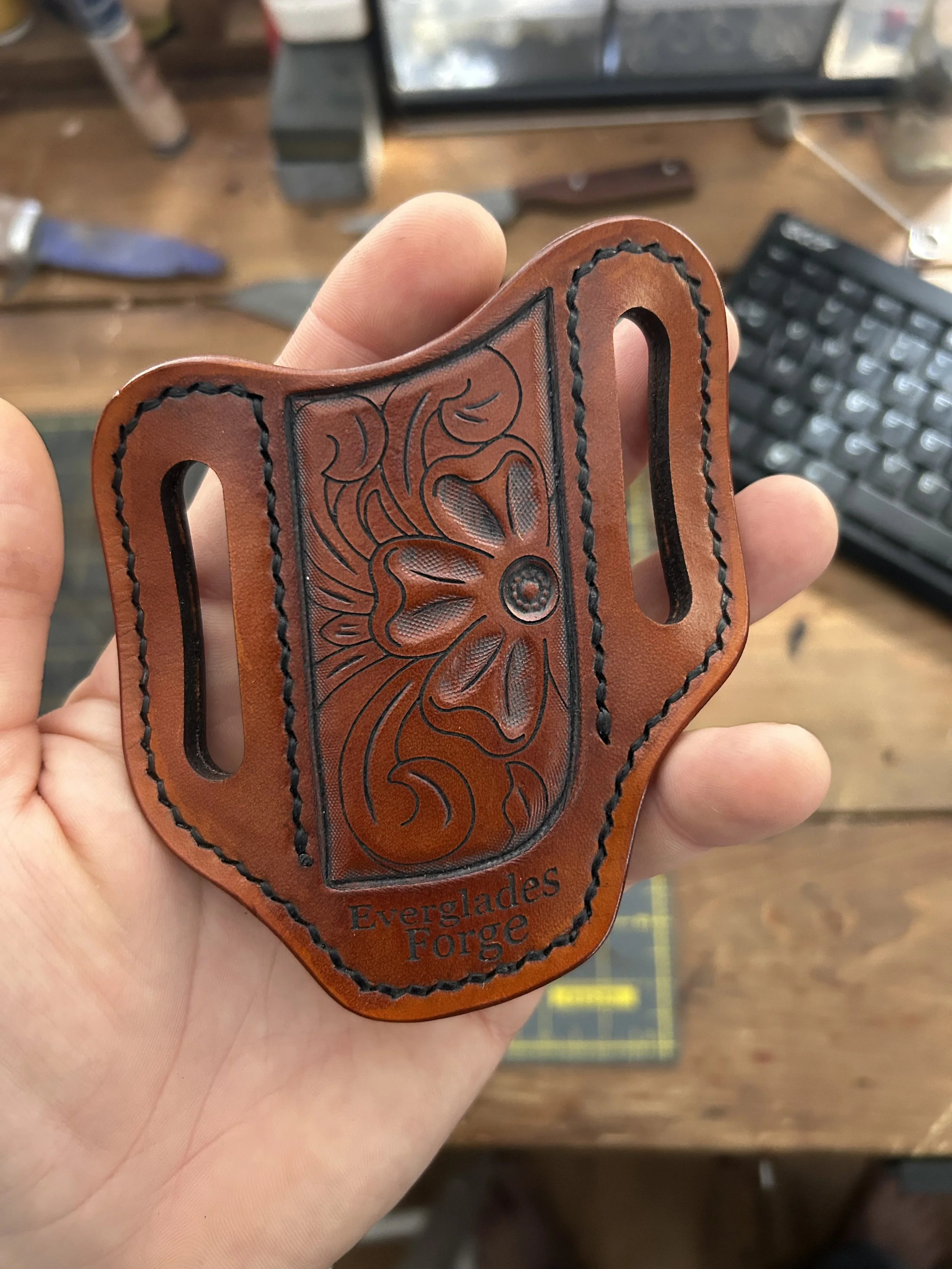 Close-up of a handcrafted leather item with engraved floral design and the words "Everglades Forge" on it, held in a person's hand over a wooden surface with a computer keyboard and some tools in the background.