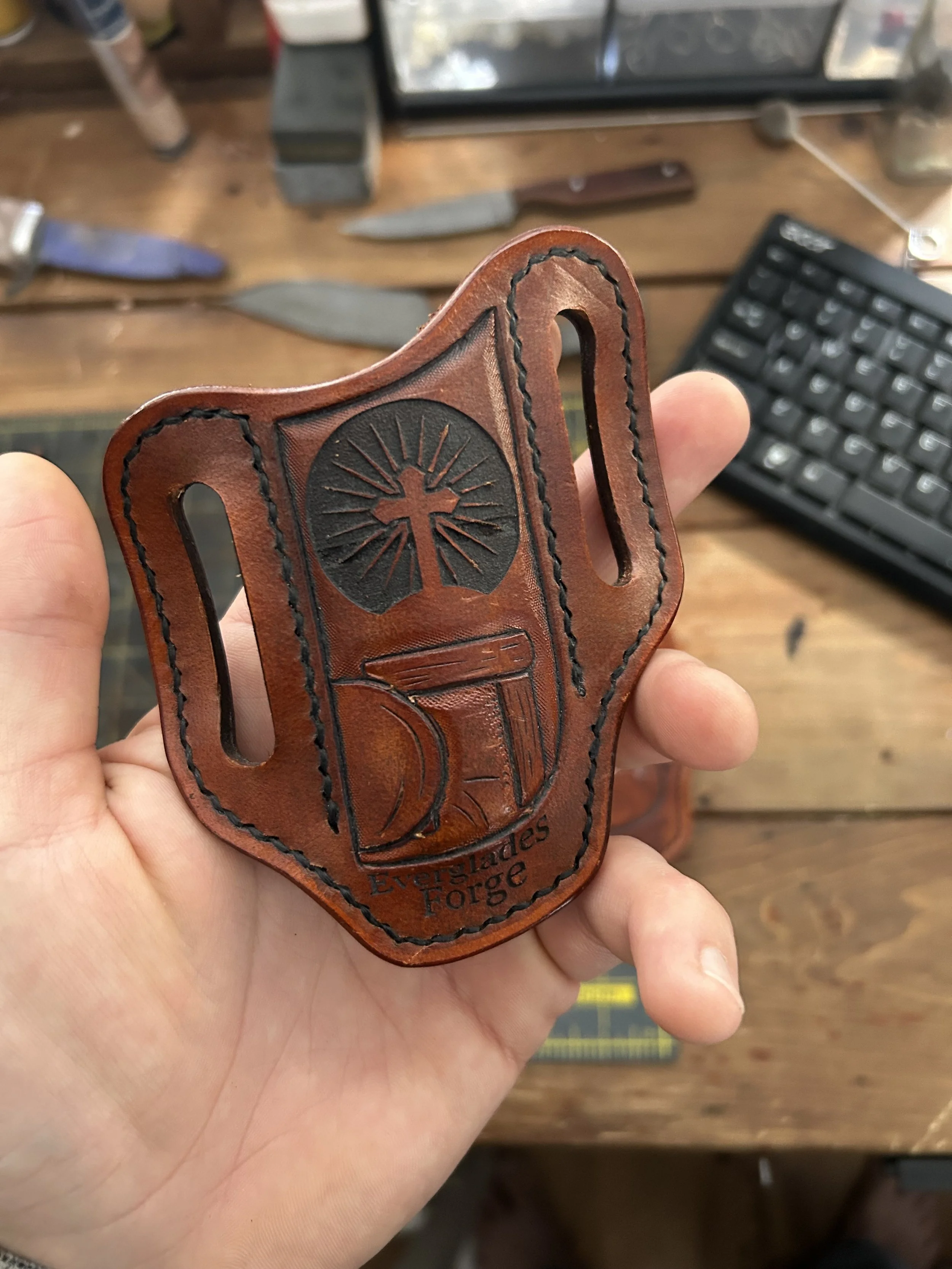 A person holding a brown leather pocket knife holster with a cross and empty tomb design, text reading 'Everglades Forge,' on a wooden workbench with knives, a keyboard, and various tools in the background.