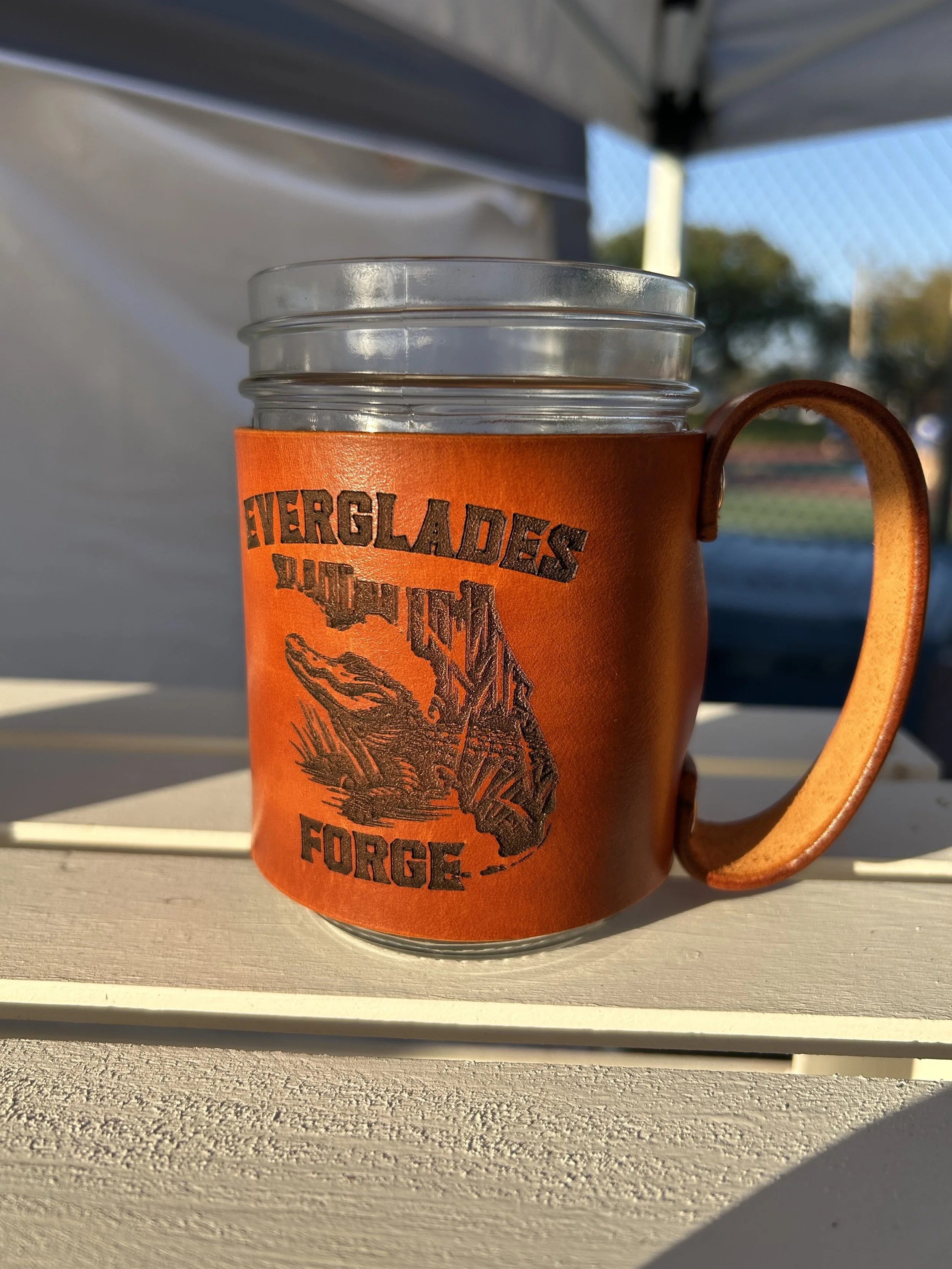 A clear glass mug with an orange leather sleeve and handle, featuring a logo that says "Everglades Outpost Wildlife" with a detailed illustration of an animal, sitting outside on a textured white surface with a blurred outdoor background.