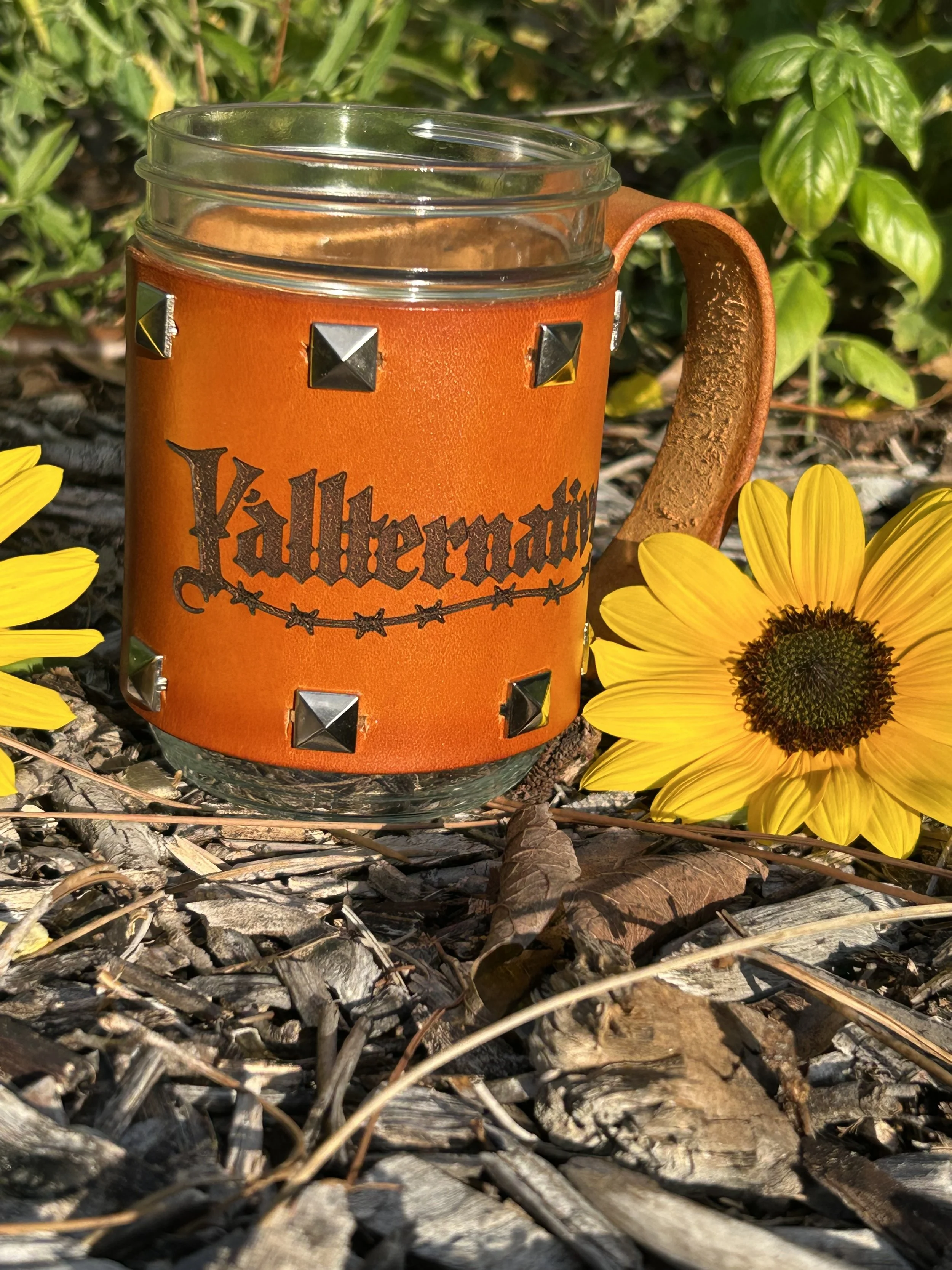 A mason jar with a leather sleeve decorated with metal studs and a label that says 'Y'allternative', surrounded by yellow flowers and fallen leaves in an outdoor setting.