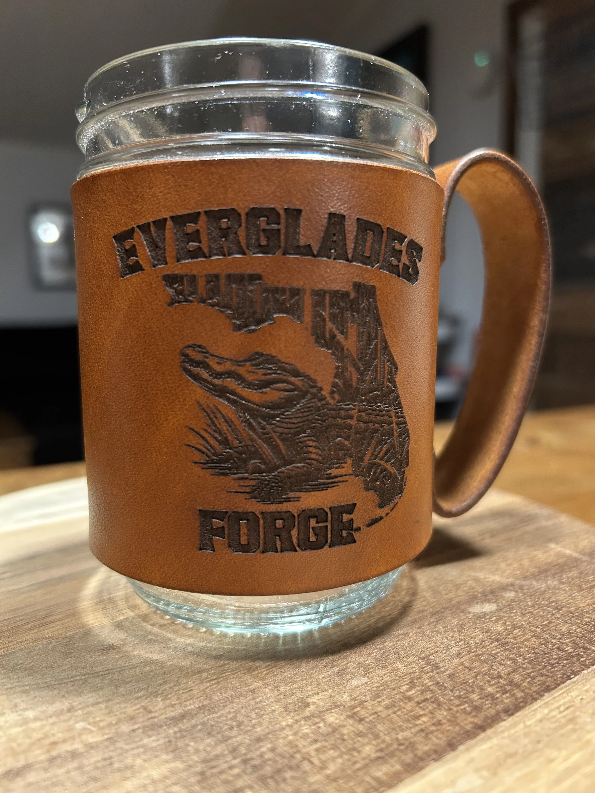 A glass mug with a leather sleeve that reads 'Everglades Motorcycle Force' and features a lizard design, sitting on a wooden surface.
