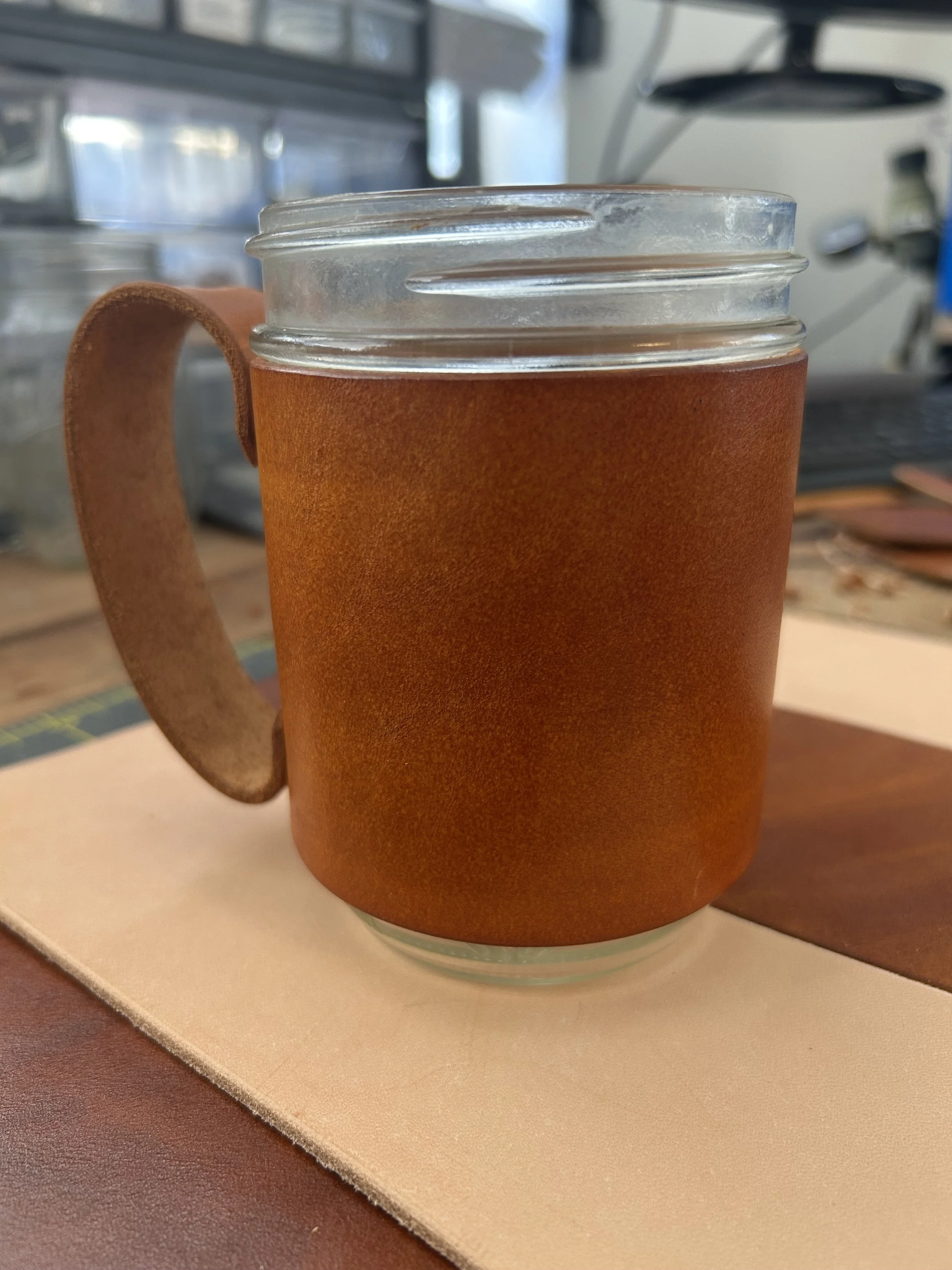 A close-up view of a glass mug with brown leather sleeve, sitting on a coaster on a desk, with a blurry background of what appears to be a computer and office equipment.