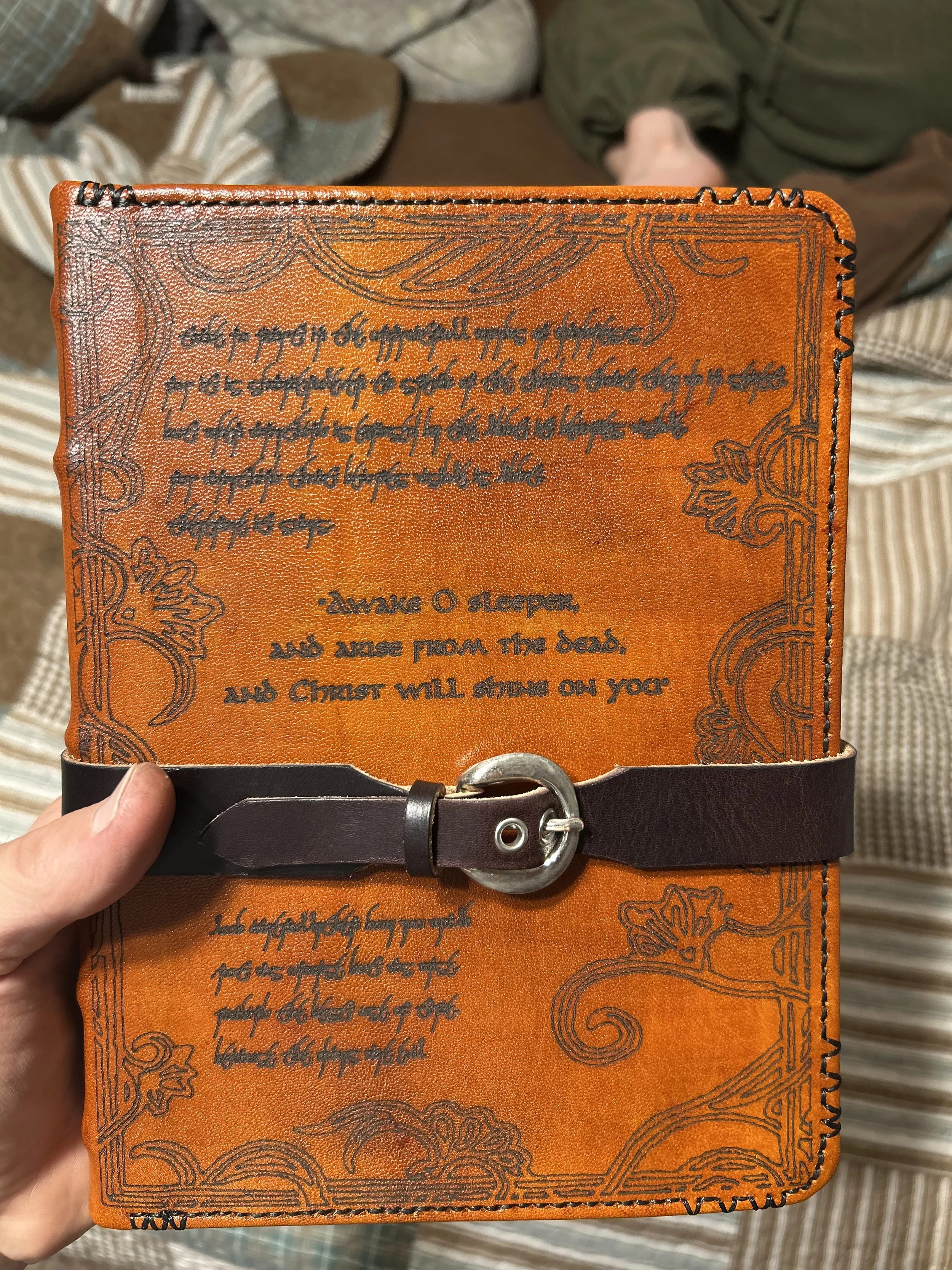 A person holding an orange book or journal with black handwritten text and decorative black border on the cover, secured with a dark brown leather strap and silver buckle. In the background, blurred shoes and clothing are visible.