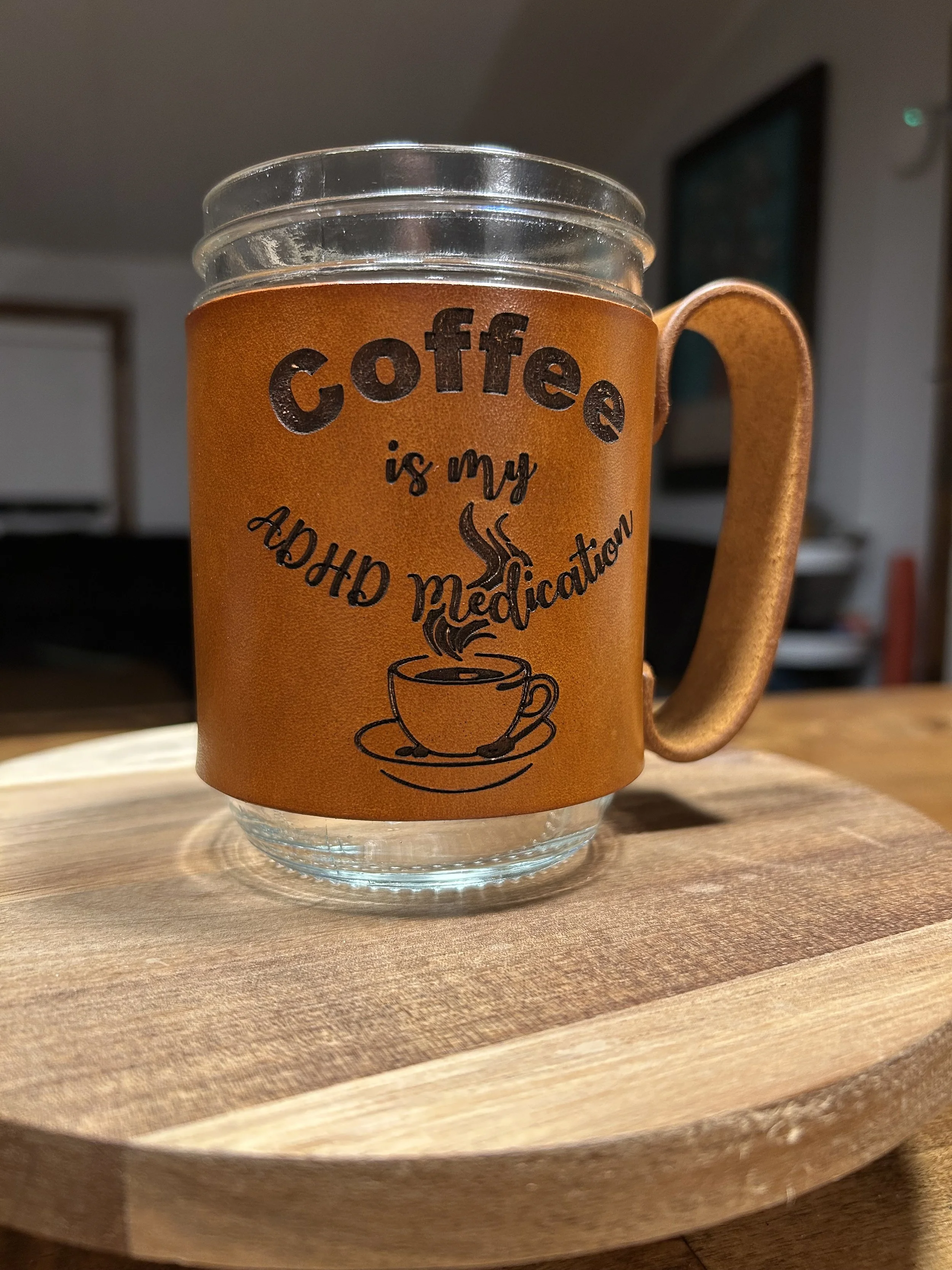 A clear glass mug with a brown sleeve and handle, featuring text and a coffee cup illustration, placed on a wooden surface.