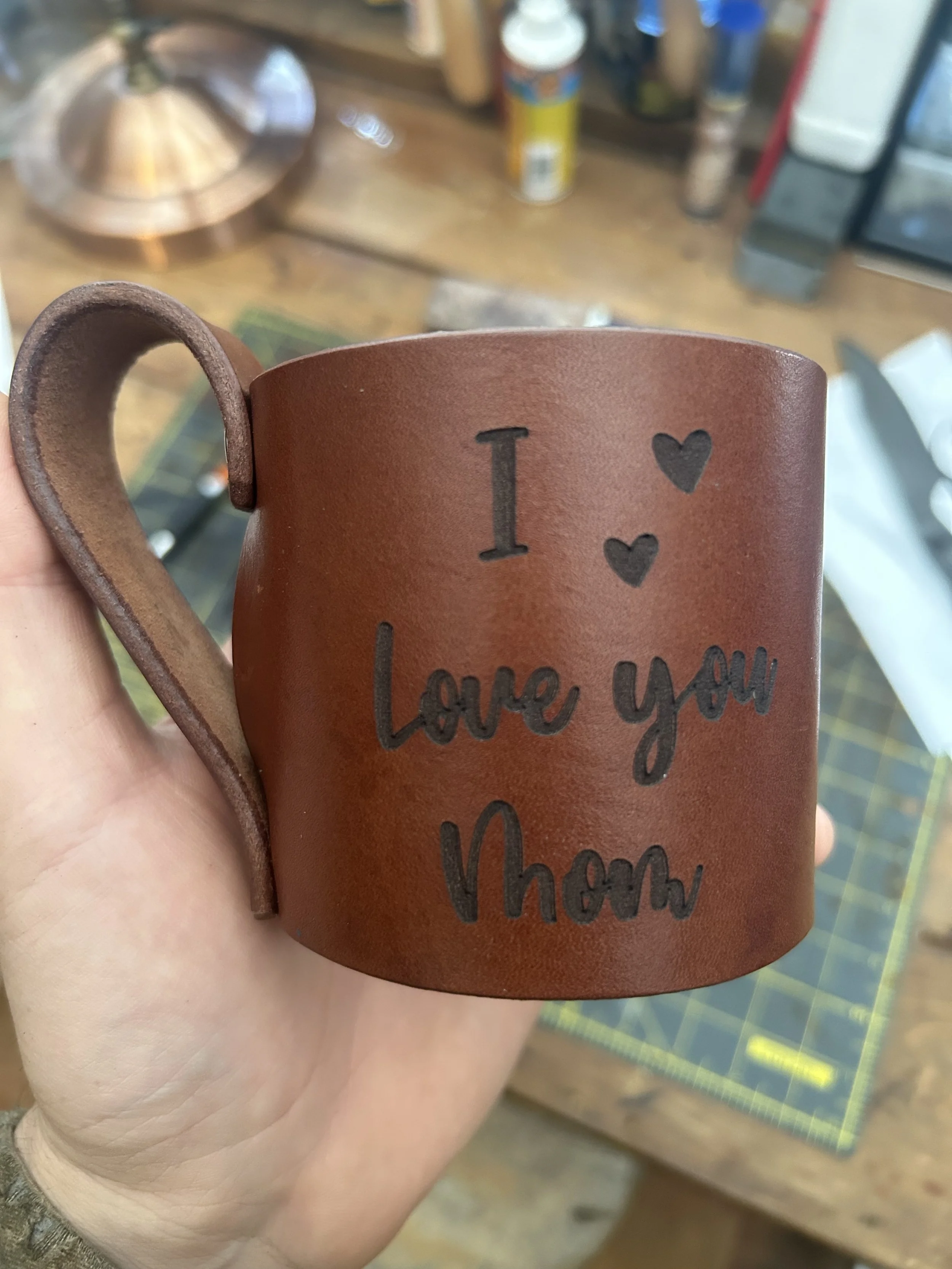 A person's hand holding a brown leather mug that says 'I love you Mom' with two hearts. The background shows a blurred wooden workbench with various supplies.