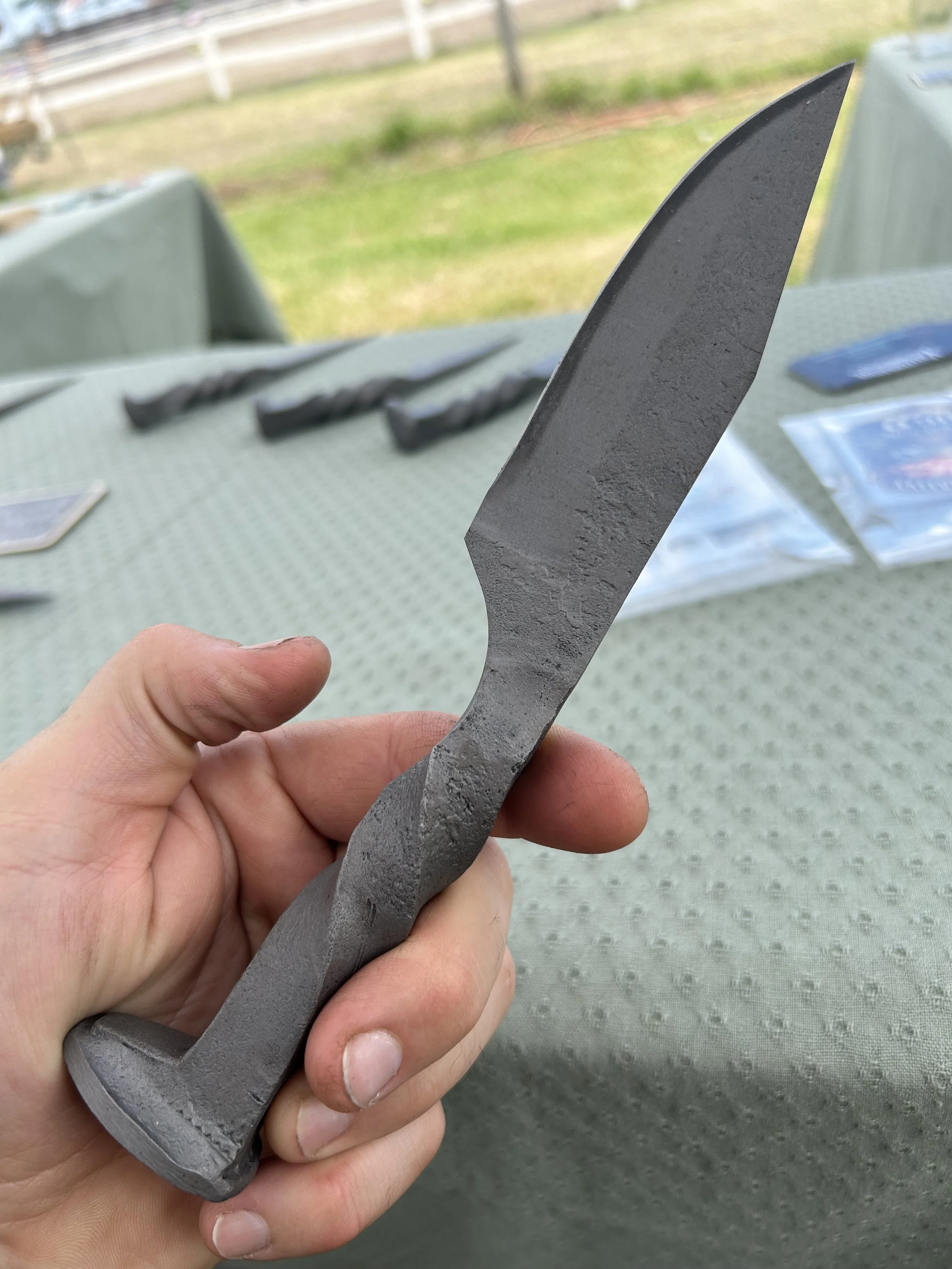 Close-up of a hand holding a gray, textured railroad spike knife with a curved blade outdoors with a blurred background of a grassy area and a wooden fence.
