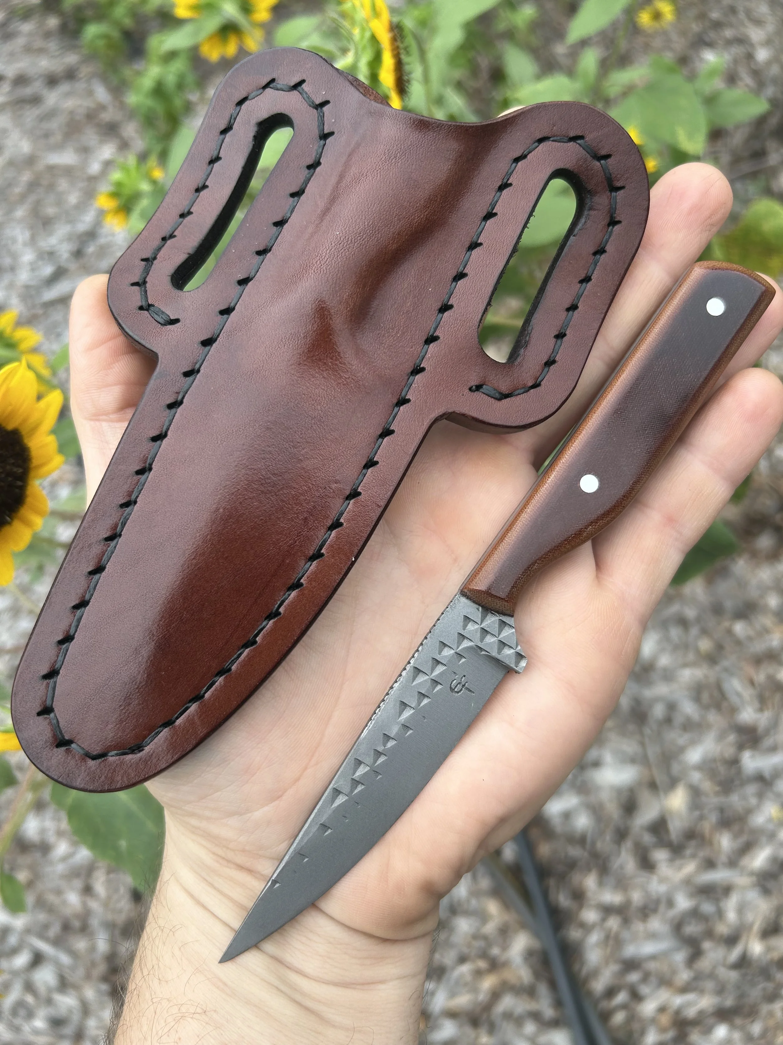 A person holding a knife with a brown leather sheath and a brown micarta handle, set outdoors with yellow flowers and green foliage in the background.