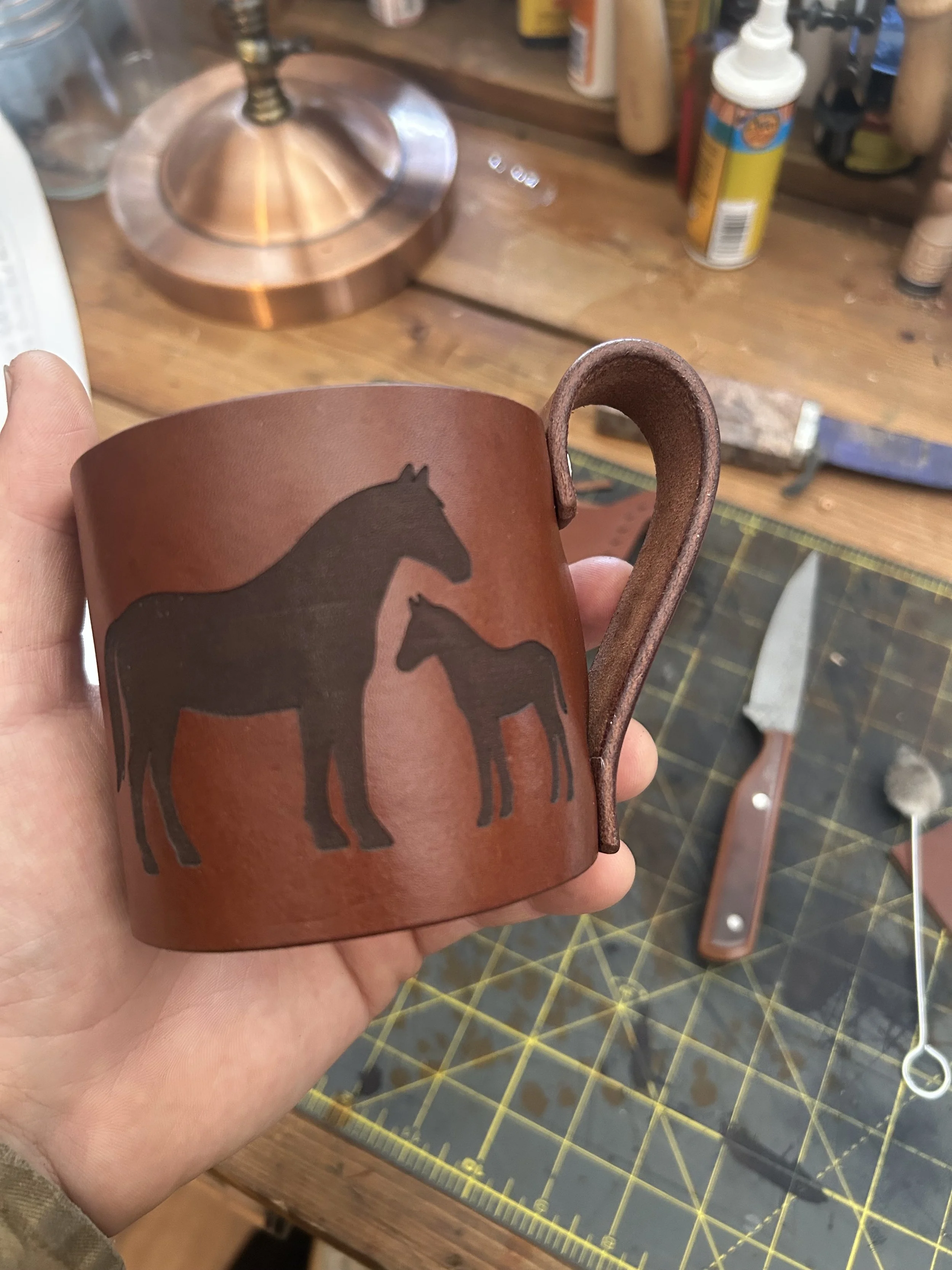 A person holding a brown ceramic mug with a black horse silhouette design, in a workshop with various tools and bottles in the background.