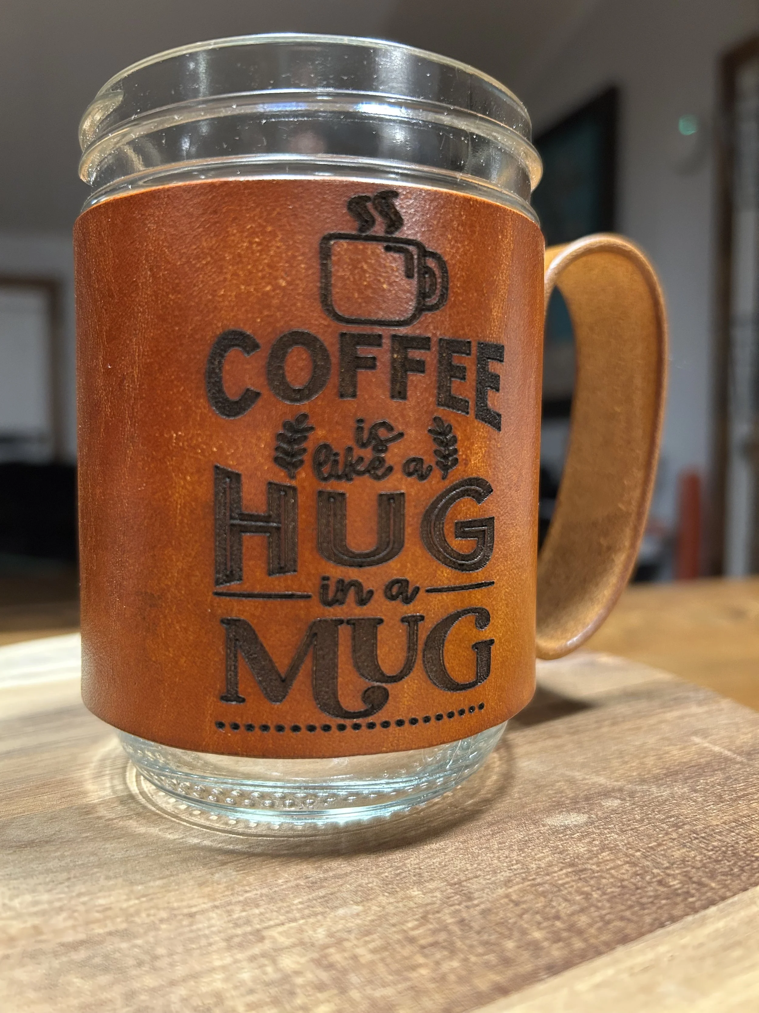 Close-up of a mason jar mug with a leather sleeve featuring a steaming coffee cup icon and the text 'Coffee is like a hug in a mug' on a wooden surface.