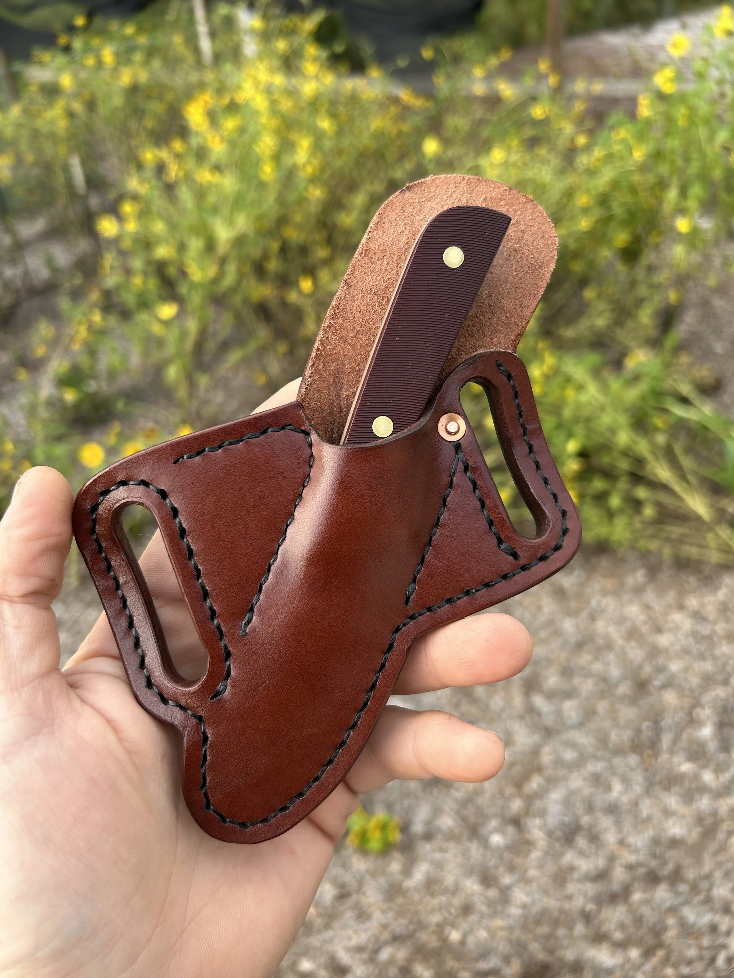 Hand holding a leather holster with a knife inside, outdoors with yellow flowers in the background.