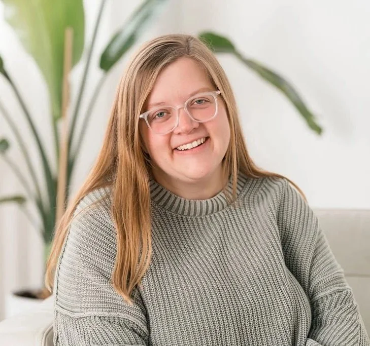 A young woman with long red hair, glasses, and a gray sweater smiling in an indoor setting with a large green plant in the background.